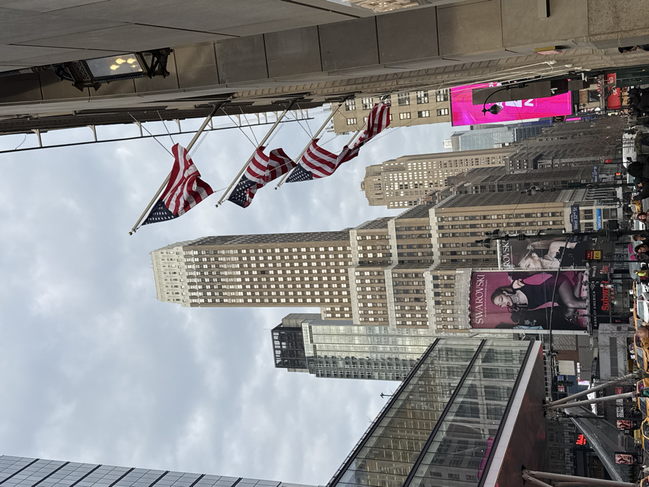 A street-level view showcases tall buildings in New York City. American flags hang from a building, with gray skies above. Billboards advertising Swarovski and Subway line the buildings. The architecture is diverse, including glass structures and older brick buildings. Pedestrians and vehicles can be seen in the background.
