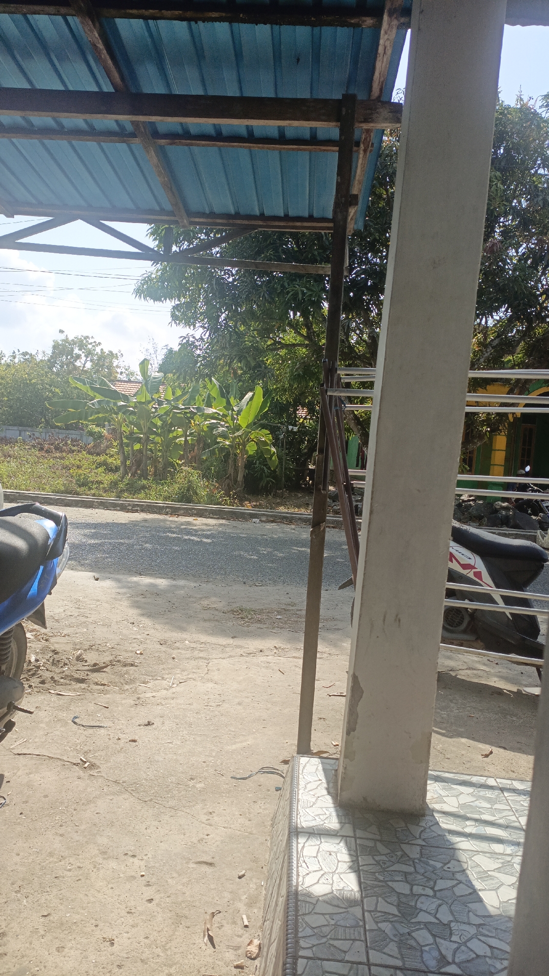 The image depicts an outdoor scene with a focus on a makeshift shelter and a street in Gusunge, Indonesia. A blue motorcycle is partially visible on the left, parked on a dirt and gravel surface. Beyond the motorcycles, a paved road runs horizontally across the frame, bordered by a low concrete curb. Across the road, a patch of land is covered in low-lying vegetation and what appear to be banana trees. Further in the background, a building with a yellow and green facade is visible, with metal fencing in front. The shelter itself is constructed with a blue corrugated metal roof supported by wooden beams and poles. A large, light-colored concrete pillar stands in the foreground on the right, obscuring a portion of the scene. The ground is a mix of dirt, gravel, and scattered debris, with patterned tiles visible at the base of the pillar. The lighting suggests a sunny day.