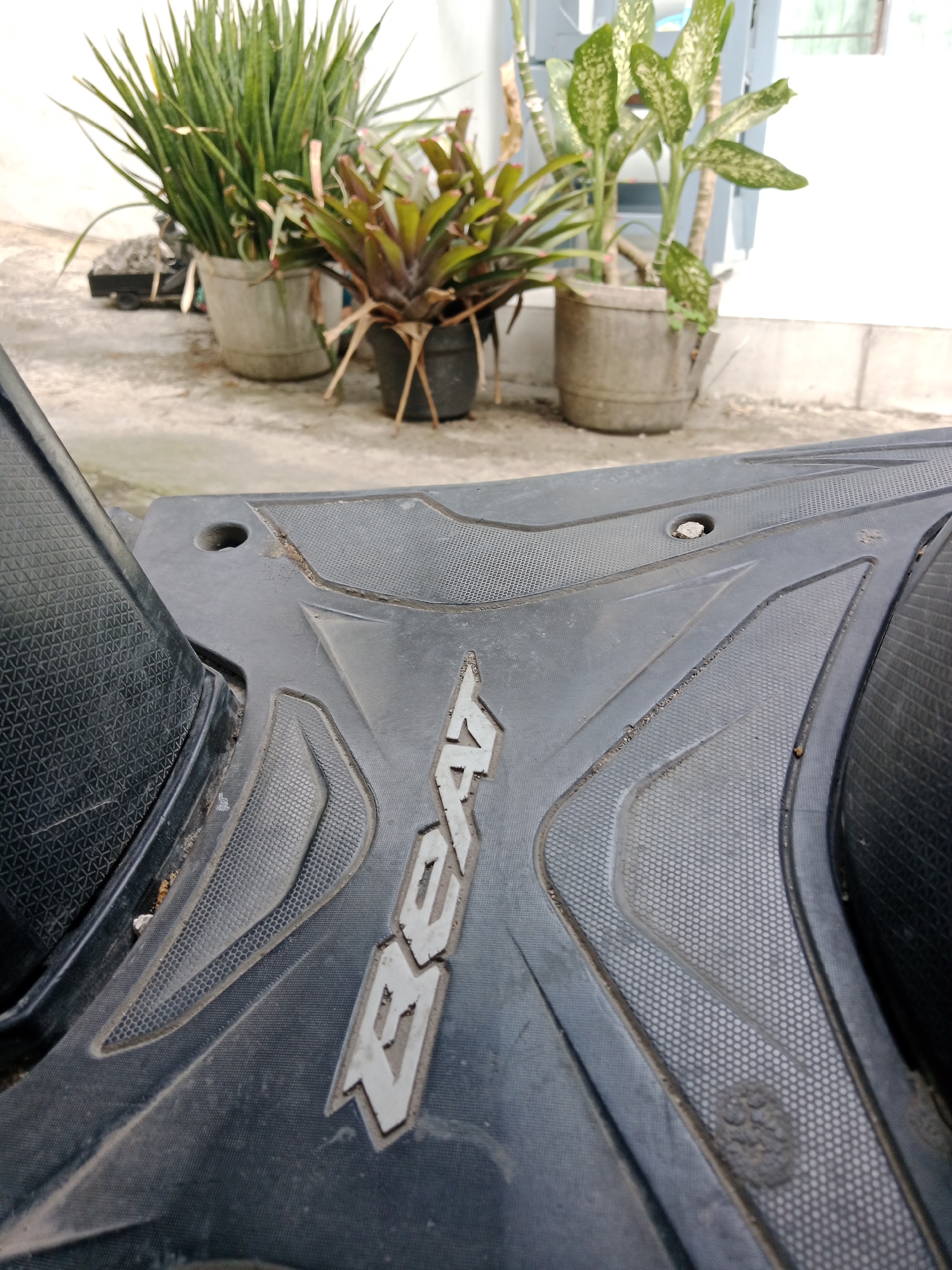 The image shows a close-up of the floorboard of a black motorcycle, with the word "BEAT" embossed in white lettering. The motorcycle floorboard is textured with a grid pattern and appears to be dusty. In the background, out of focus, are several potted plants, including a spiky green plant and a bromeliad. There are also some concrete structures visible. The scene appears to be outdoors, likely in a residential or public area, possibly a yard or a roadside. The lighting suggests it is daytime. The location is identified as Kediri City, Indonesia.