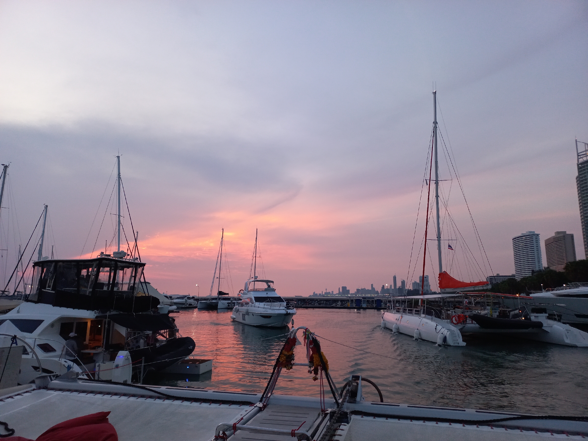 The image captures a serene marina scene at sunset or twilight. The sky is a dominant feature, displaying a striking gradient of colors from pale blue and light grey at the top to vibrant pink, orange, and red hues near the horizon, indicative of a beautiful sunset. Scattered clouds are visible, adding texture to the sky.
Numerous boats are docked in the calm waters of the marina. In the immediate foreground, the deck of a catamaran is visible, featuring a red cushion or bag. Just beyond it, another catamaran is prominent, adorned with traditional-looking colorful garlands (likely floral or fabric) hanging from its forward structure, hinting at a Southeast Asian cultural context.
To the left, a large power catamaran or yacht is moored, showing interior lights and a person in a hat visible on its deck. An inflatable dinghy is attached to its side. Various other sailboats and catamarans with their tall masts are scattered throughout the marina, many appearing as dark silhouettes against the glowing sky and water. One catamaran on the right side has a distinctive orange sail cover. The water reflects the rich colors of the sunset, creating a warm, reddish-orange sheen across its surface.
In the background, a cityscape with several tall buildings forms a silhouette against the colorful horizon, indicating an urban coastal setting. The overall mood of the scene is peaceful and tranquil, bathed in the soft, warm light of dusk. There is no visible text in the image. The location is Tambon Na Chom Thian, Thailand, which aligns with the visual cues, especially the garlands.