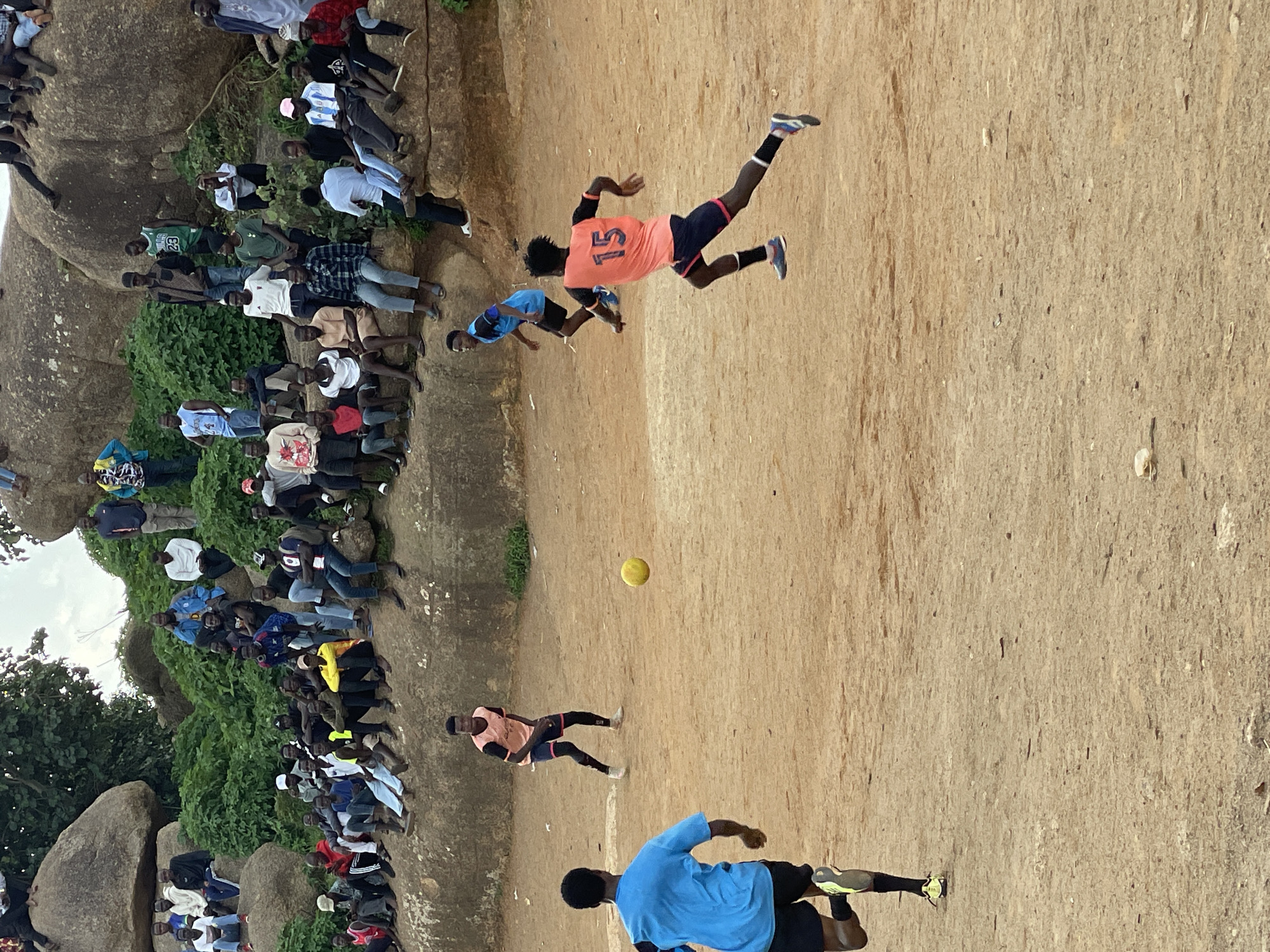 In Jos, Nigeria, a group of young men engage in a soccer match on a dusty, uneven field. The foreground is dominated by two players in action. One, wearing a bright blue shirt and black shorts, runs towards the left, his back to the camera. His opponent, in an orange jersey with the number 15 on the back, black shorts, and dark leggings, sprints to the right, his gaze fixed on the ball. A yellow soccer ball hovers in the air mid-field, a focal point of the game.
In the background, a crowd of spectators is gathered on and around large, weathered rock formations. They are seated and standing, observing the match. Their attire is casual, reflecting the warm climate. The environment appears natural and rugged, with patches of greenery interspersed among the rocks. The sky above is overcast, casting a diffused light over the scene. The overall impression is a lively community gathering centered around a spirited game of soccer.