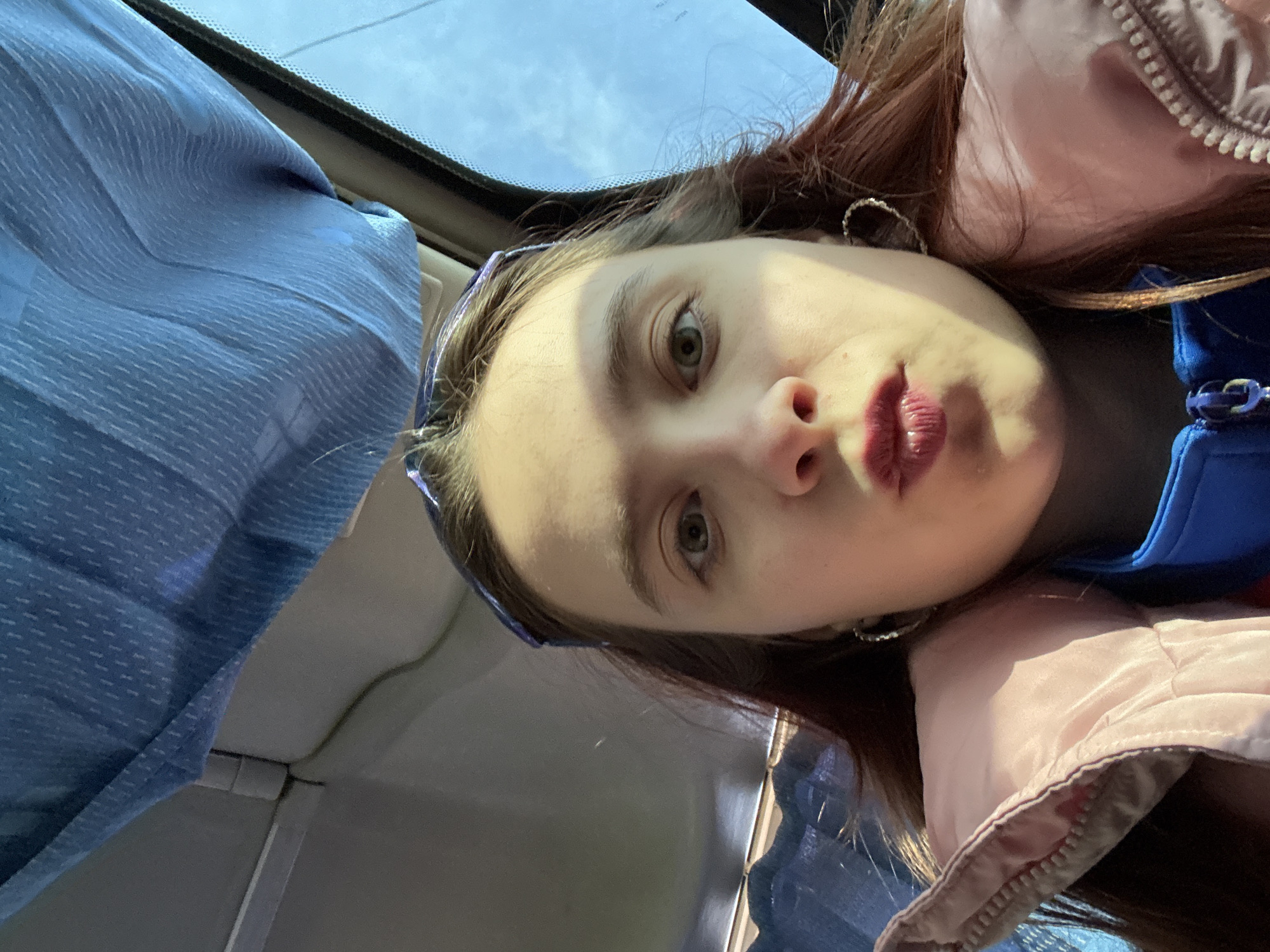 A close-up, angled selfie of a young woman with fair skin, light eyes, and auburn hair. She is wearing a blue headband and hoop earrings. Her lips are colored with red lipstick. She is wearing a pink and blue jacket. The background includes a blue curtain and what appears to be a car window with a glimpse of the sky. The location cannot be determined definitively as Berezniki, Russia, based on the given image.