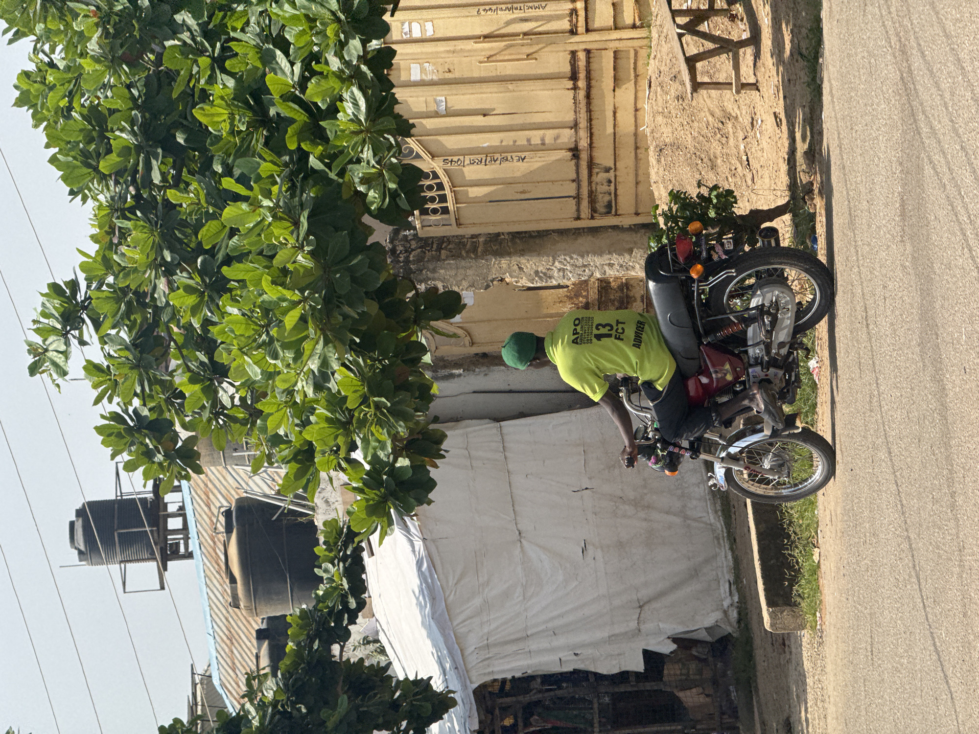 A person wearing a green cap and a bright yellow t-shirt is riding a red motorcycle down a paved road. The t-shirt has text and graphics on it, including "13 FCT ADVICER". The motorcycle has a chrome exhaust and spoked wheels.
The scene appears to be outdoors, with a large tree with green leaves in the foreground, partially obscuring a yellow corrugated metal gate with graffiti on it. Behind the gate, there is a sandy area and what looks like a tent or white tarp covering. Further in the background, there are large water tanks on top of buildings. The lighting suggests it is daytime, likely sunny given the strong shadows. The overall setting seems to be an urban or semi-urban environment in Abuja, Nigeria, with elements of informal settlements or construction visible. There are also some power lines visible. The graffiti on the gate includes what appears to be Arabic script and numbers. The text on the t-shirt is "APO KABUSA JUNCTION 07019973322 00136262709 13 FCT ADVICER".