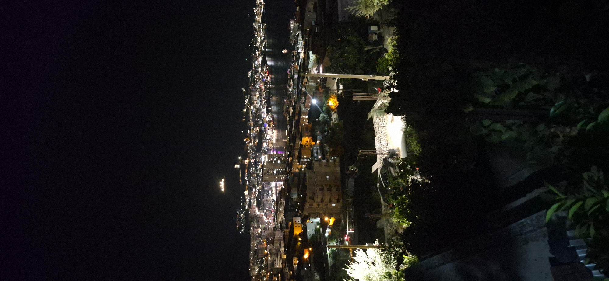 The image is a nighttime aerial view of the city of Saranda, Albania. The city lights are illuminated, reflecting on the water in the foreground. In the distance, a hill is visible with structures on top, possibly a castle or ancient ruins, lit up against the dark sky. The foreground of the image shows dark foliage and some man-made structures, including a parked car. The overall scene depicts a coastal city at night.