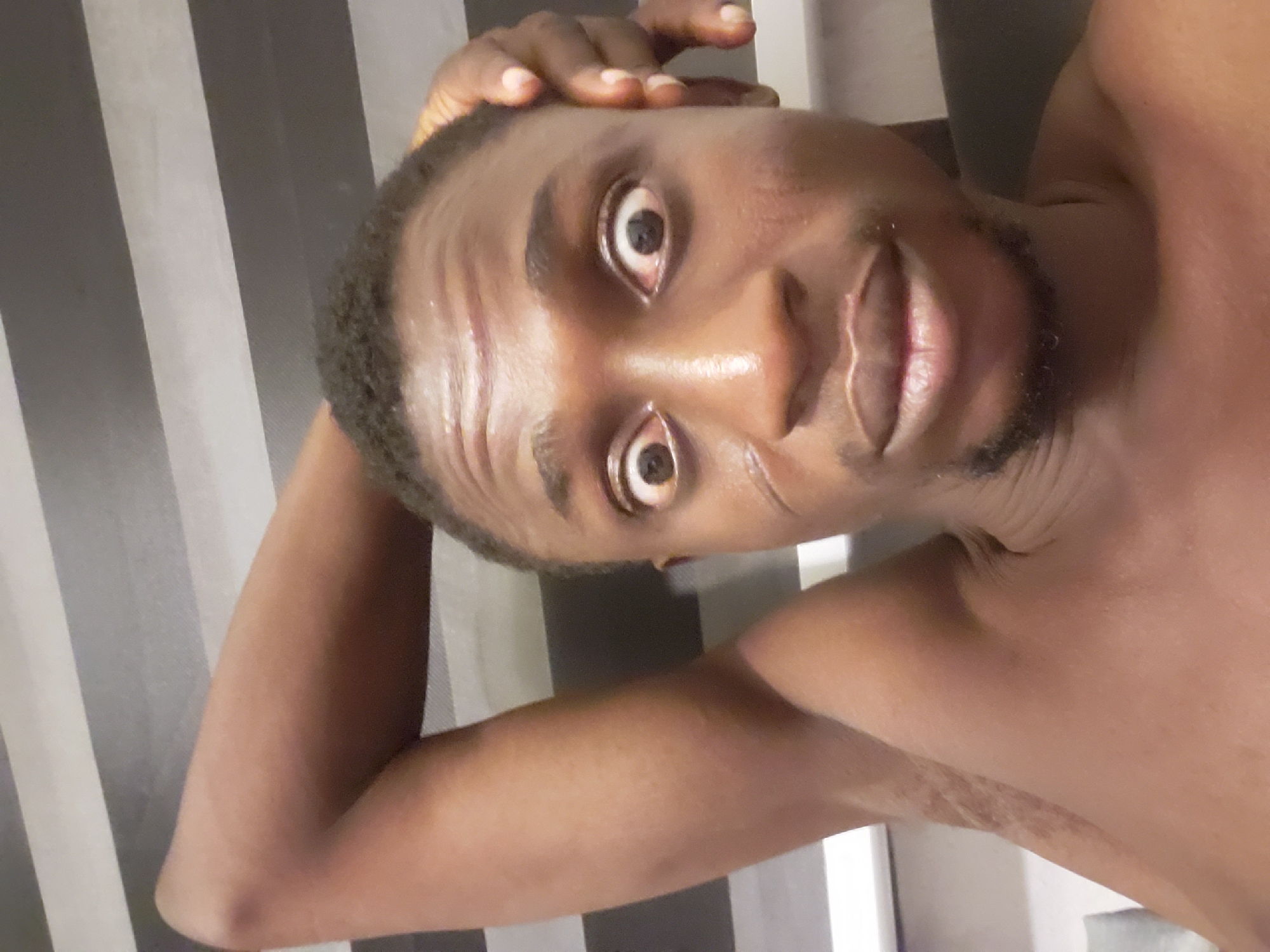 A headshot-style portrait features a dark-skinned male, likely a young adult, with short, dark, curly hair and a neatly trimmed beard or stubble. The image is rotated 90 degrees clockwise. He is looking directly into the camera with wide, exaggerated eyes, displaying prominent white sclera around his dark pupils. His mouth is slightly open, revealing pink lips. His right hand is positioned on his forehead/hairline, with fingers visible. He appears shirtless, exposing his bare shoulders and chest. His skin has a noticeable sheen, possibly from sweat, oil, or direct lighting. The background consists of distinct vertical stripes, alternating between dark gray/black and light gray/white, resembling a patterned wall or curtain. The man's expression suggests surprise, playfulness, or an intentional funny face. The photograph was taken in Adonkolo, Nigeria. There is no visible text in the image.