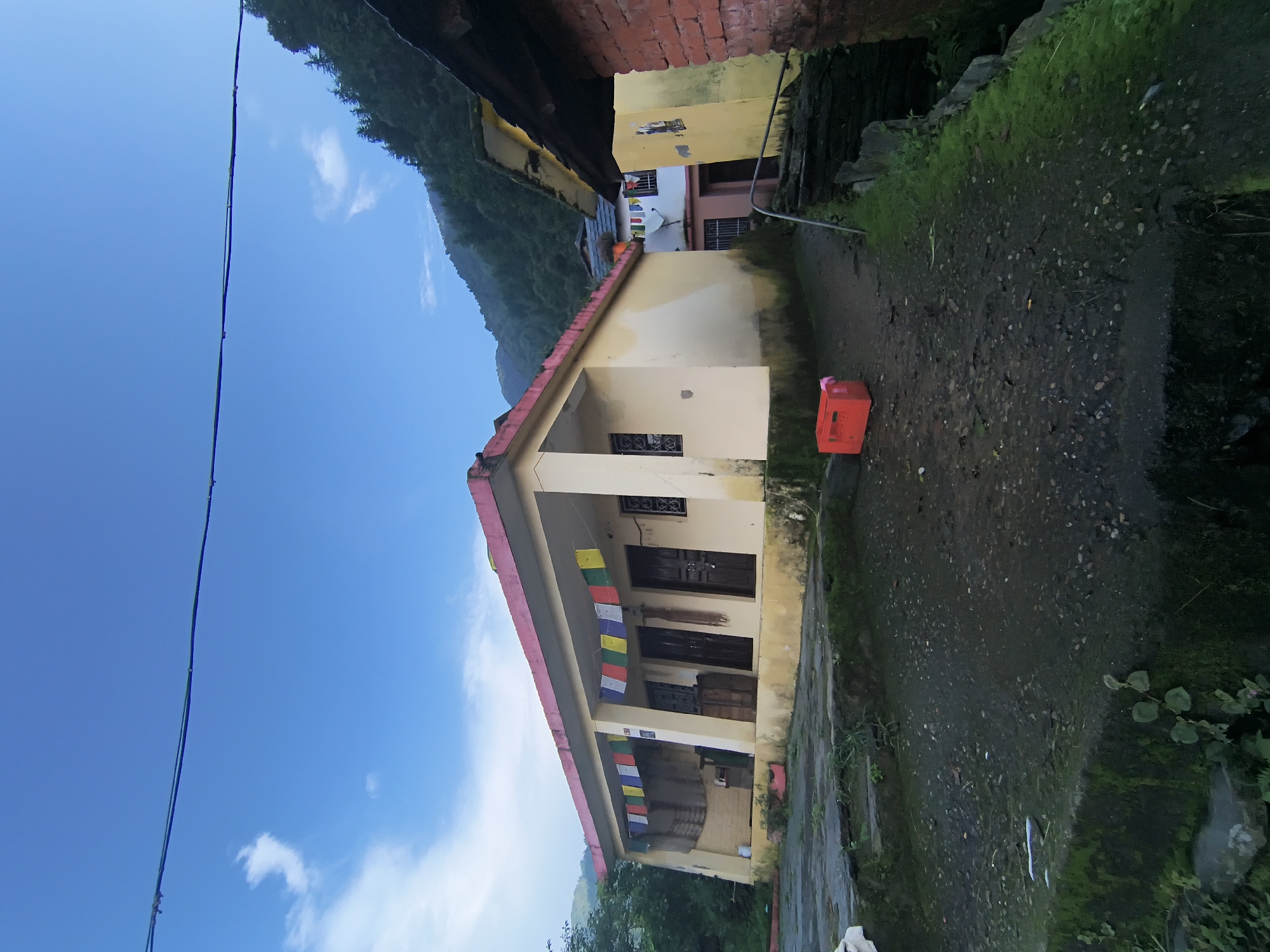 The image depicts a building in Haini, India, under a partly cloudy blue sky. The building is a single-story structure with a yellow exterior and a pink trim along the roofline. Prayer flags in various colors are hung across the front porch. The ground in front of the building is paved with dark stones and is partially covered in moss and greenery. To the right of the main building, a pathway leads between it and another building constructed of red brick. In the foreground, a bright red plastic crate sits on the ground. Beyond the buildings, a lush green hillside rises, covered in trees.