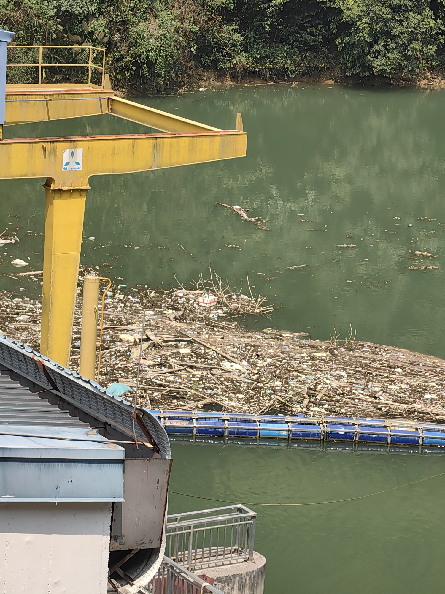 The image shows a body of water heavily polluted with debris, including plastic and wood. A yellow metal structure stands above the water. A blue boom is visible, attempting to contain the trash. Part of a building, with a gray roof and walls, is in the foreground. The location is Bảo Lạc, Vietnam.