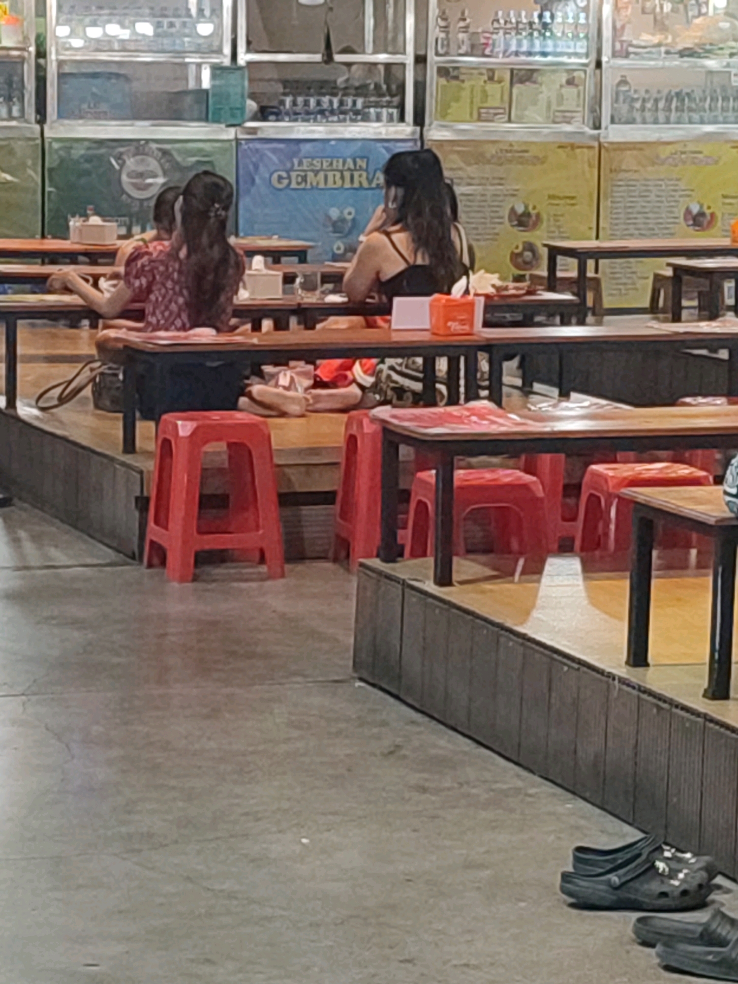 The image depicts an outdoor seating area of a food establishment in Yogyakarta, Indonesia. In the foreground, a concrete floor shows some cracks and a pair of black Crocs and black slides are placed on the ground. A wooden partition with vertical slats forms a barrier. Behind this, a tiered seating area features several red plastic stools and wooden tables. Two women are seated at one of the tables in the mid-ground. One woman, facing away from the camera, has long dark hair and is wearing a black tank top. The other woman, also facing away, has dark hair tied back and is wearing a patterned red and white top. They are sitting at a wooden table with a white tissue box and an orange container on it. In the background, various shelves are stocked with bottles, and colorful banners hang on the wall. One banner displays the text "LESEHAN GEMBIRA". The overall setting suggests a casual dining experience, likely at a lesehan (floor seating) restaurant common in Indonesia.