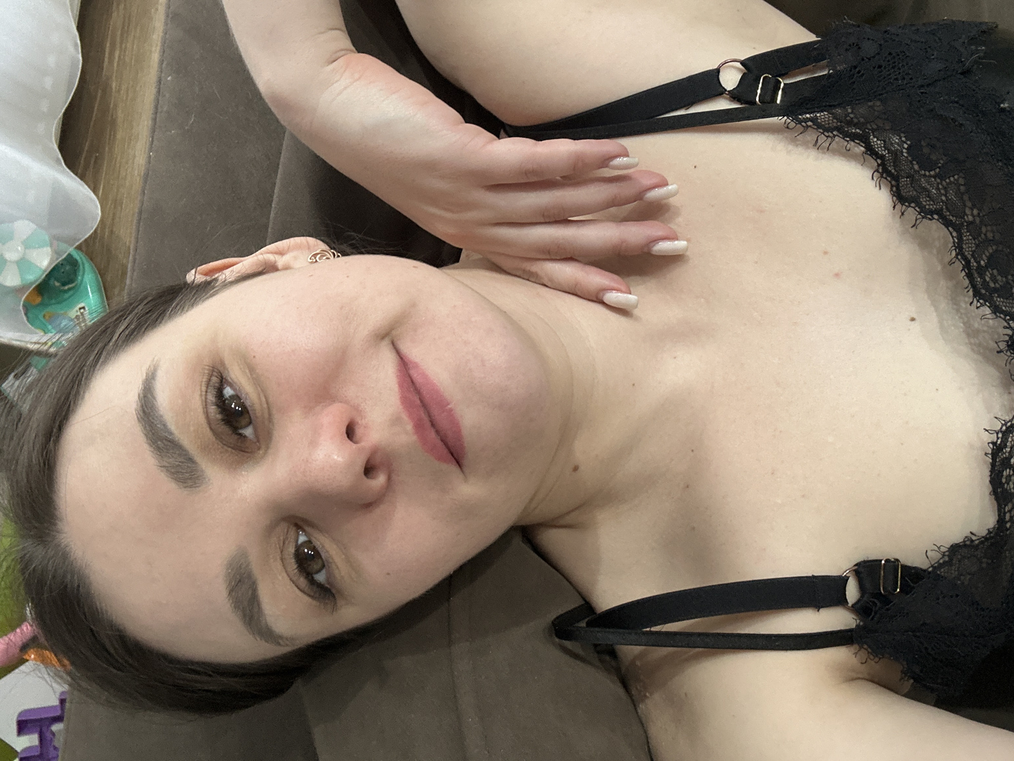 An eye-level shot captures a young woman lying on her back, her head turned to face the camera with a gentle, slightly coy smile. Her dark hair is pulled back, framing a face with well-defined dark eyebrows and expressive dark eyes. Her lips are painted a muted rose shade. She is wearing a black lace top or bra with thin straps and metallic gold-colored rings and adjusters visible on her right shoulder. Her left arm is raised, with her hand resting delicately near her collarbone, showcasing long, manicured nails with a light, possibly glittery polish. Her skin appears fair and unblemished. She lies on a dark brown surface, likely a sofa or bed. In the background, particularly in the top and bottom left corners, out-of-focus household items are visible, including what appears to be a light-colored plastic object, a green container, and some colorful, possibly child-related toys. The lighting is soft and even, suggesting an indoor setting during the daytime or under gentle artificial light. The overall mood is relaxed and intimate. No visible text is present in the image. The location is Chaikovsky, Russia.