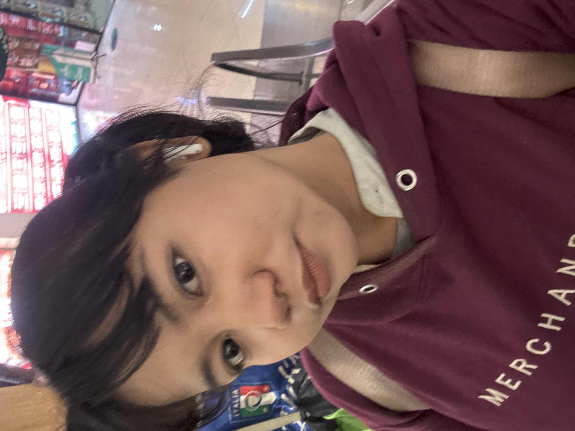 A close-up selfie of a young woman with dark hair pulled back into two pigtails, wearing a maroon hoodie and a light-colored shirt underneath. She has dark eyes and is wearing white earbuds. The text "MERCHAND" is visible on the front of her hoodie. In the background, an "ITALIA" logo is partially visible on a blue garment. The environment appears to be indoors, possibly a shopping mall or public space, with a tiled floor and metal railings visible. The lighting is bright and appears to be artificial. The image does not contain any information that would allow for specific location identification within North Jakarta, Indonesia.