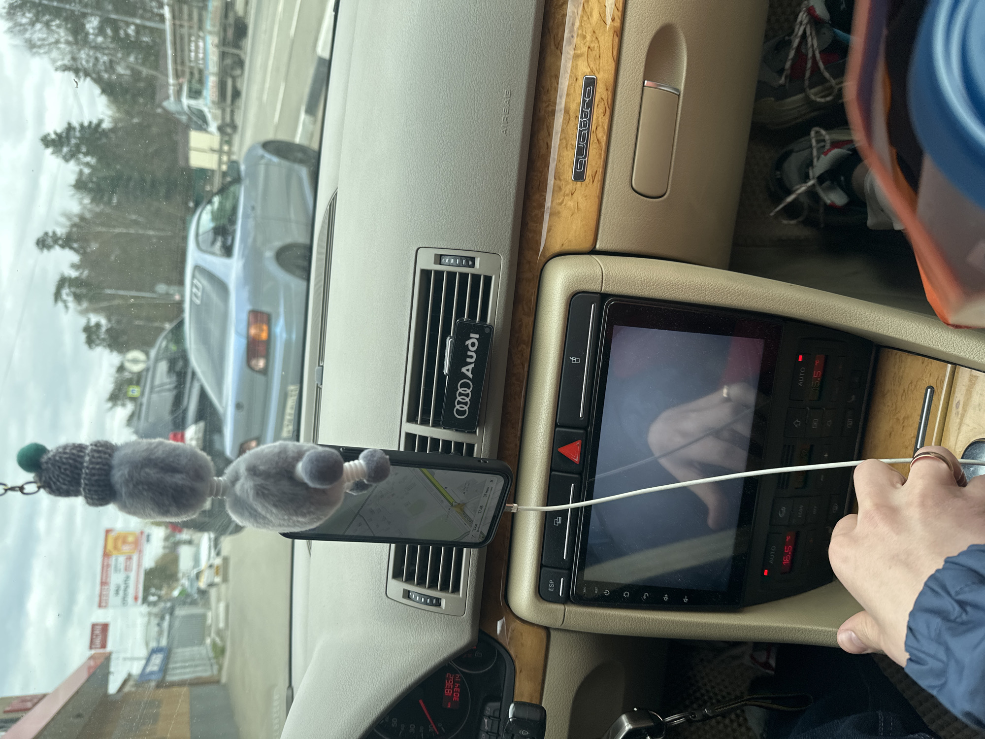 The image provides a view inside an Audi car, likely an older model, from the passenger's perspective, looking towards the dashboard, central console, and the road ahead. The car features a wooden dashboard trim with "Quattro" branding and an "AIRBAG" label above the glove compartment.
A person's right hand, adorned with a rose gold ring, is visible on the right, interacting with the car's central controls. The car's climate control system is integrated into a modern touchscreen display, showing "AUTO" mode and a temperature setting of "16.5" degrees Celsius, with "15.5" also visible. Buttons for functions like "ECON" and "ESP" are present below the screen. A white charging cable connects to a smartphone.
The smartphone is mounted on a central air vent, displaying a map interface indicating the time "17:45" and a remaining travel time of "38 мин". Another air vent features an "Audi" branded air freshener. Part of the speedometer and odometer are visible in the lower left, showing readings of "1836.2" and "3034.14". A decorative grey fuzzy air freshener with a knitted cap hangs from the rearview mirror.
In the passenger's footwell or storage area, a pair of sneakers, possibly Balenciaga Triple S, are stored along with a blue cup or container.
Through the windshield, an overcast sky and a street scene typical of Malakhovka, Russia, are visible, featuring other vehicles, trees, and buildings, indicating the car is on the road or in traffic.