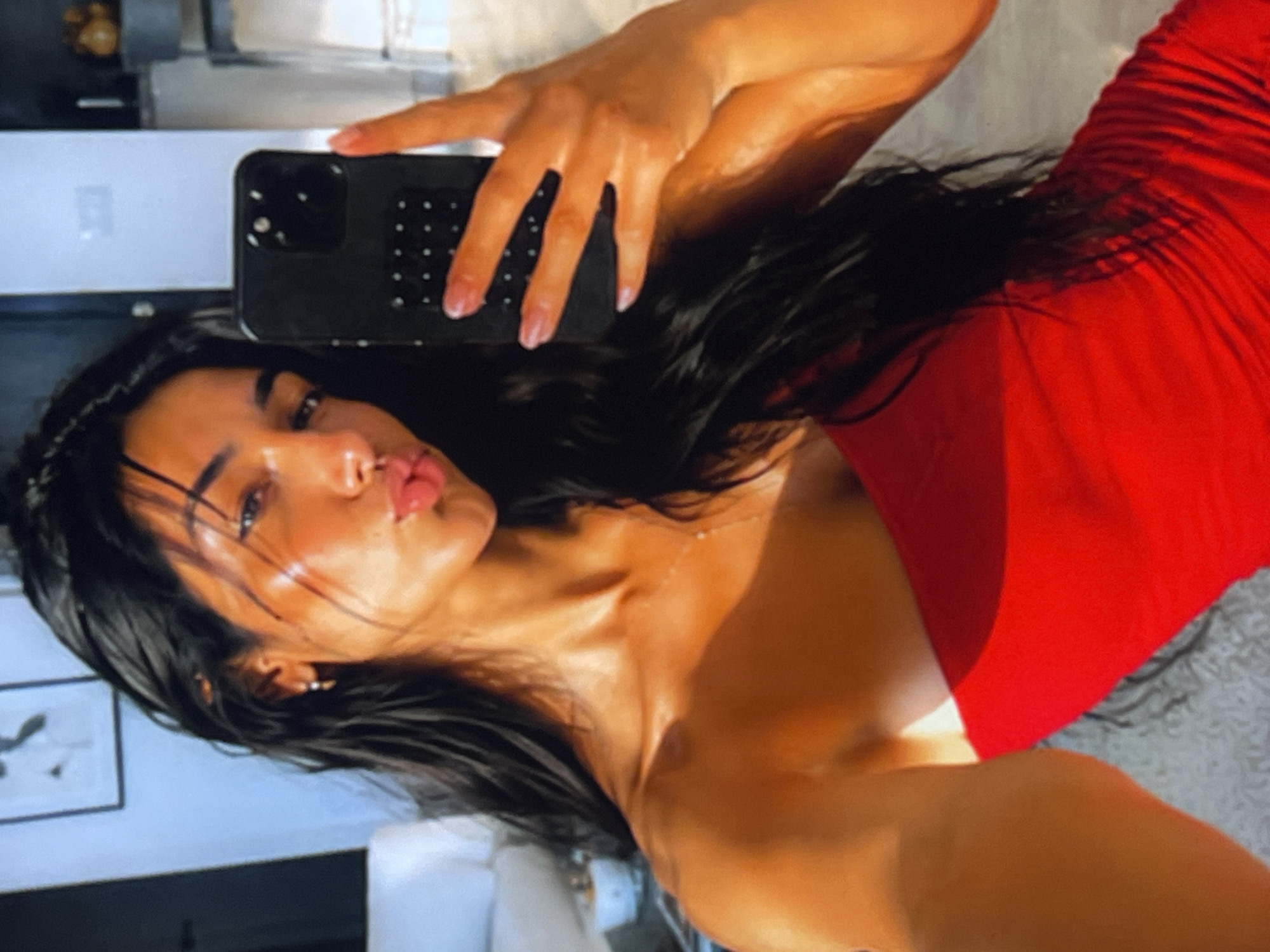The image is a vertical, close-up selfie of a woman wearing a red dress. She is holding a black smartphone in her right hand, which is angled to capture her face and upper body. The woman has long, dark hair that falls around her shoulders and is slightly damp. Her skin appears dewy and illuminated, suggesting a warm, possibly artificial, light source. She is looking at the camera with her lips pursed in a kissing gesture, and her eyes are partially closed. Her expression conveys a sense of playful sensuality or a posed moment for social media.
The background is blurred and indistinct, but hints of a domestic setting can be observed. To the left, a framed picture is partially visible, appearing to be a sketch or drawing. Behind the woman's head, there's a hint of a wall or furniture with a reflective surface. The overall lighting and composition suggest the photo was taken indoors, possibly in a bedroom or living area, at a time when artificial lights are on, as natural daylight does not seem to be the primary source of illumination. The focus is entirely on the woman and her selfie, with the background serving only to provide a minimal context.