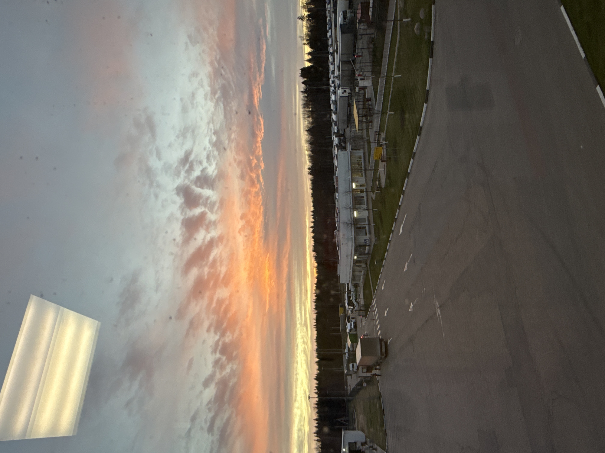The image shows a view from a high vantage point in Lupanovo, Russia. A large expanse of gray asphalt road with directional arrows stretches into the distance. To the right of the road is a strip of grass with various metal poles and equipment. Beyond the grass is a row of white, rectangular buildings. A line of trees borders the buildings. A colorful sunset or sunrise fills the sky on the left side of the image, featuring shades of orange, pink, and white clouds. In the lower left corner, there's a rectangular, reflective surface that appears to be artificially lit. The angle of the shot suggests a building or elevated structure as the origin point of the photograph.