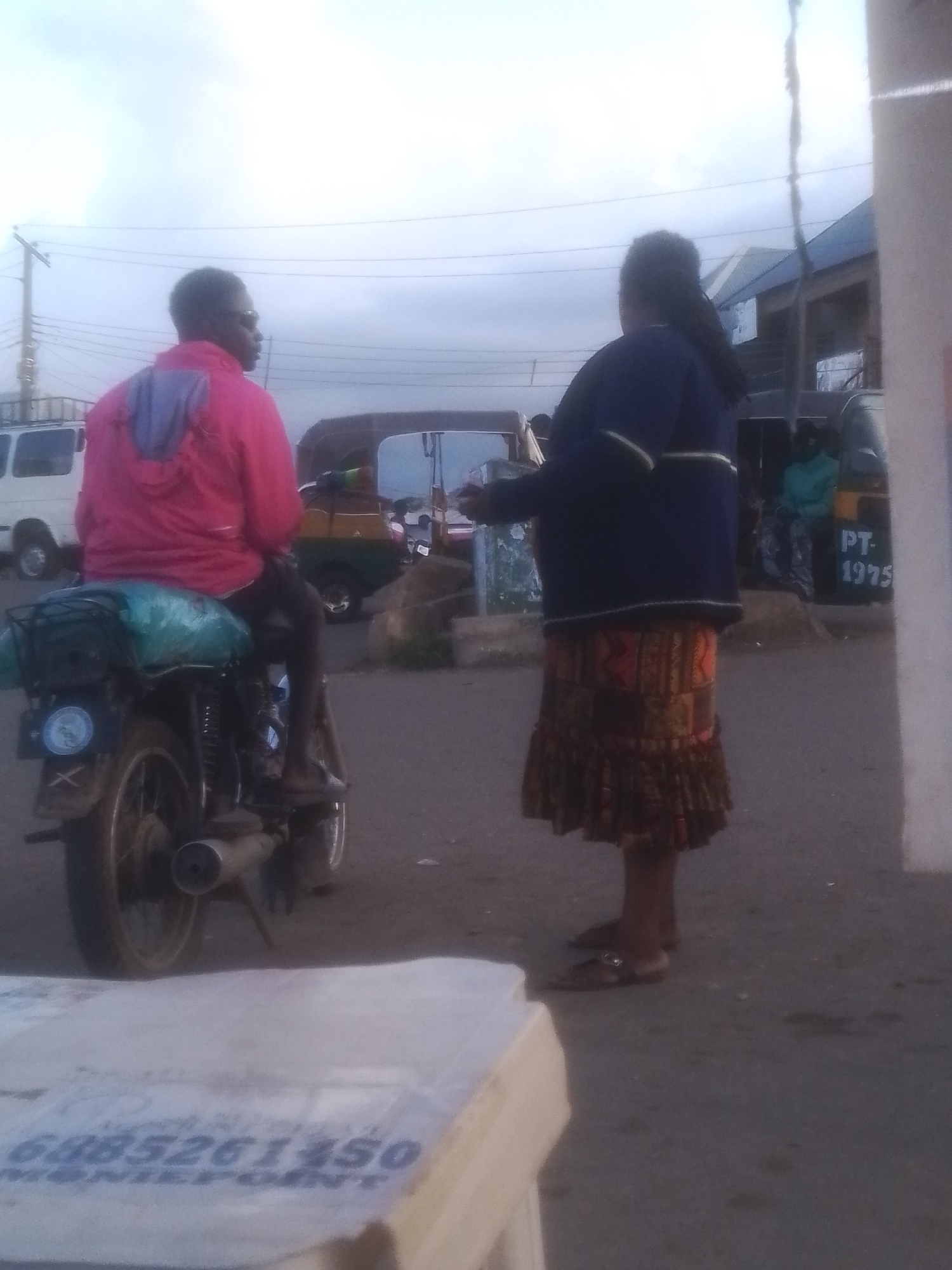 A low-angle, slightly blurry, medium shot depicts two individuals standing on a paved road, possibly in Gwong, Nigeria. The person on the left, viewed from the back and partially obscured by a motorcycle, is seated on the motorcycle wearing a vibrant pink hooded jacket over darker clothing. The motorcycle is dark-colored, loaded with items wrapped in a light blue tarp on its rear seat and rack. The individual on the right stands facing left, seen from the back and right side, wearing a dark blue jacket with white stripes on the sleeves and a patterned, calf-length skirt in earthy tones. They are wearing sandals. The background reveals a street scene with a white van parked to the left and a yellow and green three-wheeled auto-rickshaw with "PT-1975" written on its side in the distance to the right. Other vehicles and structures, including a building with a blue roof, are visible further in the background. The sky is overcast. In the foreground, a portion of a white, rectangular object with some visible text is present.