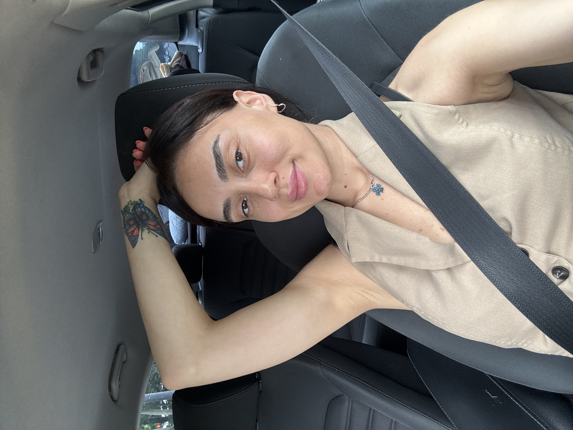 A woman is captured in a selfie taken from inside a car. She is seated in the driver's seat, wearing a light beige sleeveless vest and a necklace with a cross-shaped pendant. Her right arm is raised and resting behind her head, revealing a butterfly tattoo on her upper arm. The car's black seatbelt is prominently visible across her chest. The background shows the interior of the car, including the headrest, and a glimpse of an outdoor environment through the car window, suggesting the car is in motion or parked in a city. The specific location of Tbilisi, Georgia, is not discernible from the visual information in the image.