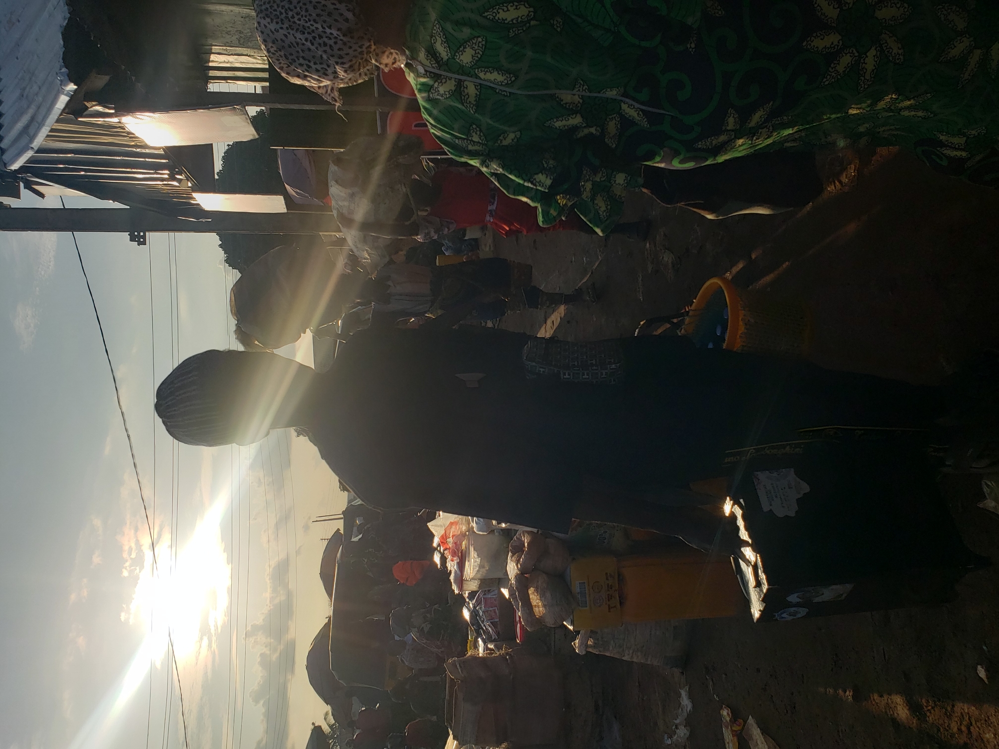 In Kakuri Gwari, Nigeria, a market scene unfolds under a bright, sunlit sky with visible sun flares. A person, silhouetted by the sun, stands in the foreground carrying a dark suitcase. To the right, another person is visible, wearing a green patterned garment. The background reveals a bustling marketplace with tents, market stalls, and various goods, including sacks of produce and crates. The ground appears to be dirt. The overall atmosphere suggests daytime activity within a busy, open-air market.