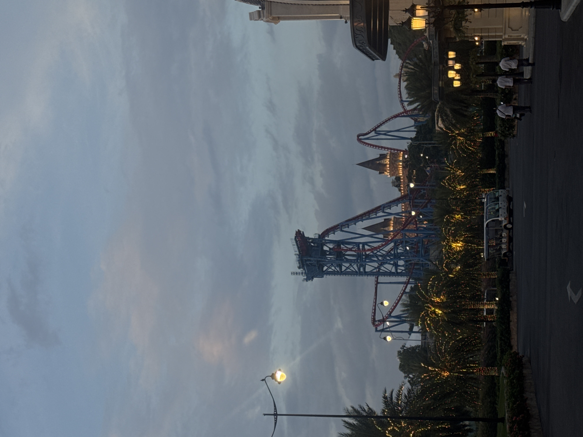 An outdoor, medium-shot, vertical image shows the entrance area of a theme park at dusk. The sky is cloudy and a dusky blue. A large roller coaster with blue tracks and red supports dominates the background, with two castle-like structures visible behind it, illuminated by warm lights.
In the foreground, a paved pathway leads towards the theme park. On the left, a tall lamppost with a yellow light stands beside a line of palm trees decorated with numerous small, twinkling lights. On the right, a building facade with ornate architectural details and several warm yellow lamps is visible. Three people are walking away from the camera down the pathway towards the right side of the frame. Two small white vehicles, similar to golf carts, are parked on the pathway between the palm trees and the building. The scene suggests the VinWonders Nha Trang amusement park on Hon Tre Island, off the coast of Nha Trang, Vietnam.