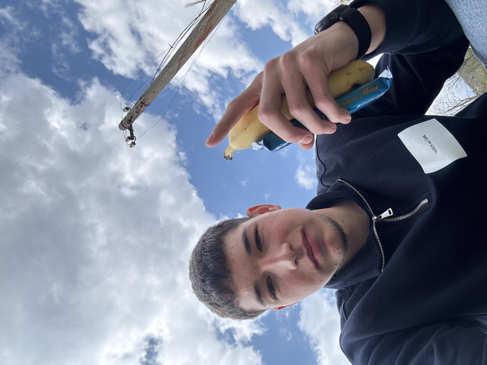A low-angle selfie taken outdoors with a partly cloudy sky. A power pole is visible in the upper left of the frame. A person holds a banana and a wrapped snack bar ("Den u Hou"). The person is wearing a black jacket, a black watch, and a white patch with non-English text is visible on the jacket. The location is Dobromirtsi, Bulgaria.