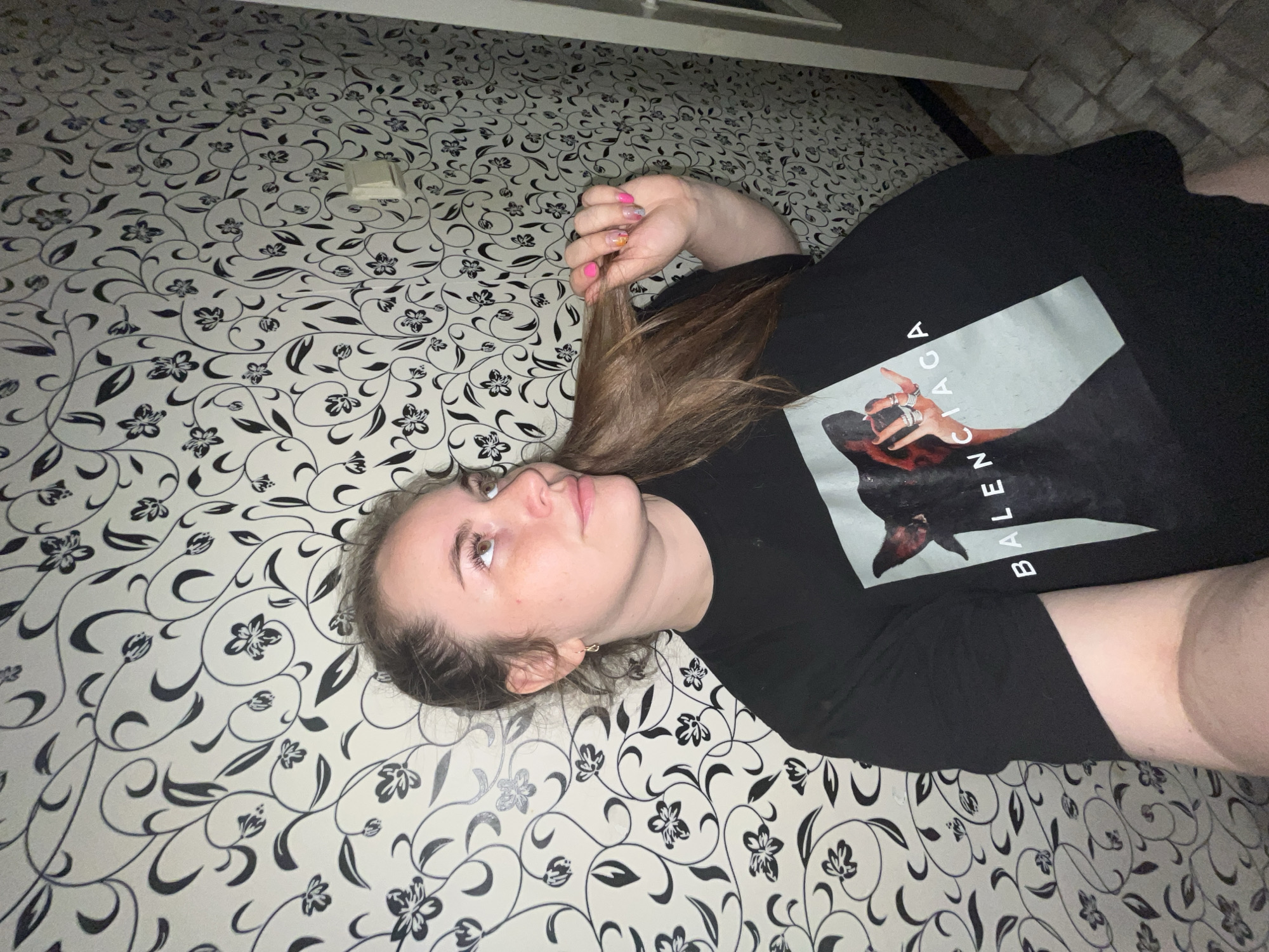 A young woman is lying on a surface with a black and white floral pattern, possibly wallpaper or a bedspread. She is wearing a black Balenciaga t-shirt. Her hair is light brown and extends towards her outstretched hand, which has painted nails. She is looking slightly upward and to the side. The image appears to be taken indoors, but there is no indication of the location being in Chaikovsky, Russia. The environment includes a neutral wall with some texture in the background and a white edge that might be a window frame or part of a piece of furniture.