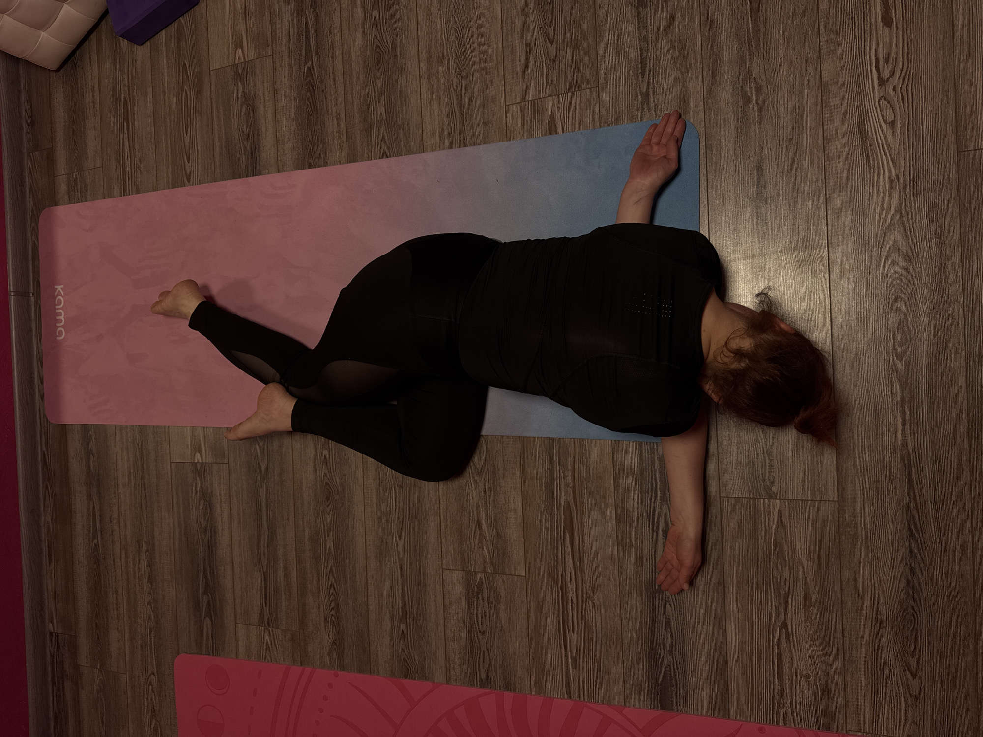 The image depicts a person lying on a yoga mat in a supine spinal twist pose. The yoga mat is pink and blue and has the word "KAMA" on it. The person is wearing black leggings and a black top. The person's body is twisted to the left, with the legs bent and knees stacked. The arms are extended to the sides. The person's head is turned to the right. The floor is wooden. The location is likely indoors. There is no other evidence to identify the location as the city of Chaikovsky, Russia.