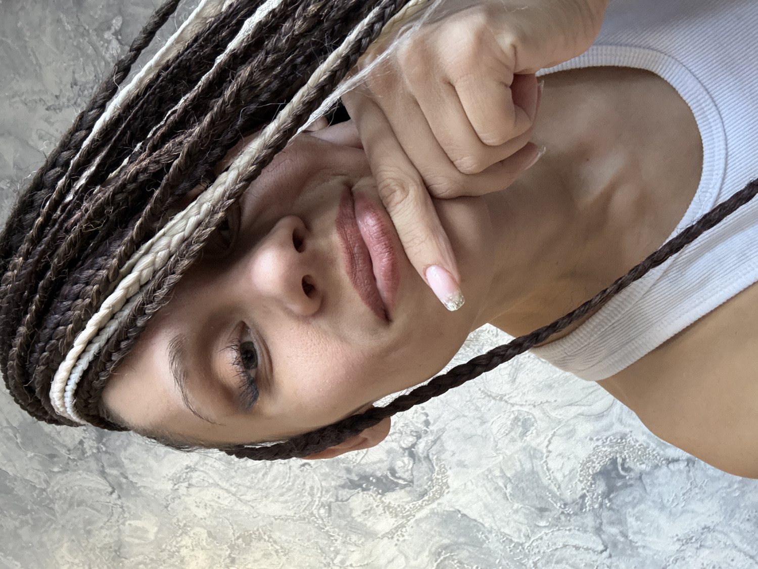 A close-up, rotated portrait features a person, likely a woman, with their head tilted, looking directly at the viewer. The subject's face shows a subtle, pensive expression with slightly pursed lips. Their hair is styled in numerous long, fine braids, predominantly dark brown with noticeable strands of white or very light blonde interwoven throughout, framing the side of their face. A hand, with long, manicured fingernails adorned with a light pink base and sparkling silver tips, rests gently against their jawline. The person is wearing a simple white ribbed tank top, visible around the neck and shoulder area. The background is a light-colored, textured surface with varying shades of grey and white, suggesting an indoor setting. There is no visible text in the image. This casual portrait was captured in Rusanovo, Russia.