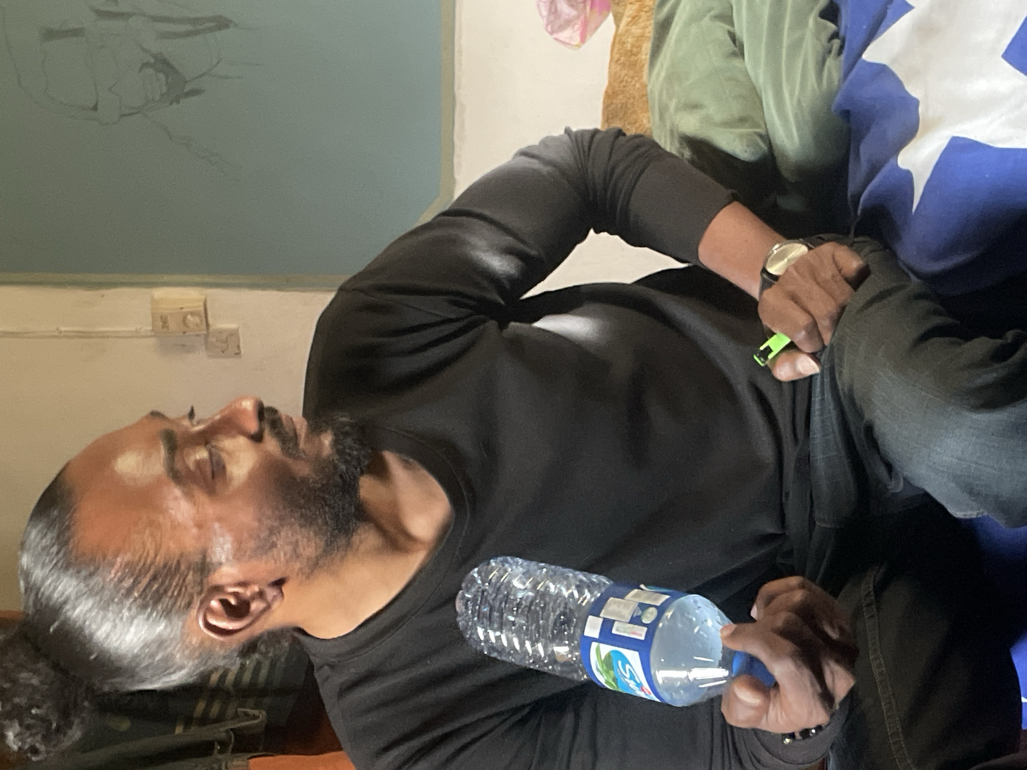 A medium indoor shot depicts a dark-skinned man with long hair pulled back and a full beard, wearing a long-sleeved black shirt. He's reclining and holding a plastic water bottle. On his left wrist, he sports a wristwatch with a light-colored face. His right hand is also visible, holding a small green object. The background features a light blue wall with electrical outlets and what appears to be peeling paint near the ceiling. A blanket or garment with a blue and white star pattern is situated on the right side of the frame, along with a green garment. The location is likely indoors. There is no information to verify its exact location as Jos, Nigeria.
