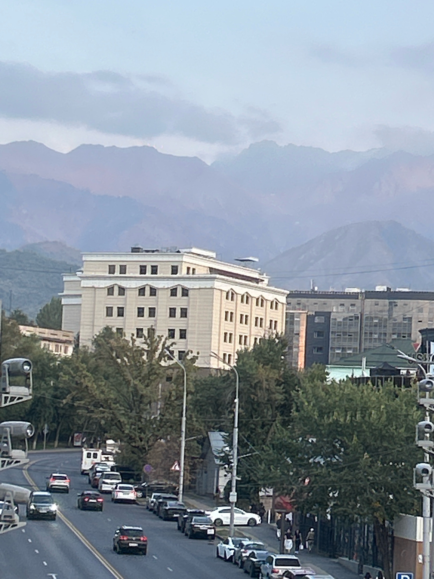 A medium shot captures an urban landscape with a prominent building and a backdrop of mountains. In the foreground, a road curves to the left, with several cars driving on it. Some of the cars are parked along the side of the road. Trees line the road and the area around the buildings.
In the middle ground, a light-colored, multi-story building stands out, characterized by its symmetrical design and numerous windows. To its right, a darker, more modern building with glass facades is visible. Further back, more buildings and trees create a dense urban environment.
The background is dominated by a range of hazy, purple-hued mountains under a pale sky. The lighting suggests it is either early morning or late afternoon. The overall scene depicts a view in the city of Almaty, Kazakhstan, where the urban environment meets the natural grandeur of the mountains. There are traffic lights and cameras present along the road. No people are clearly distinguishable in the image.