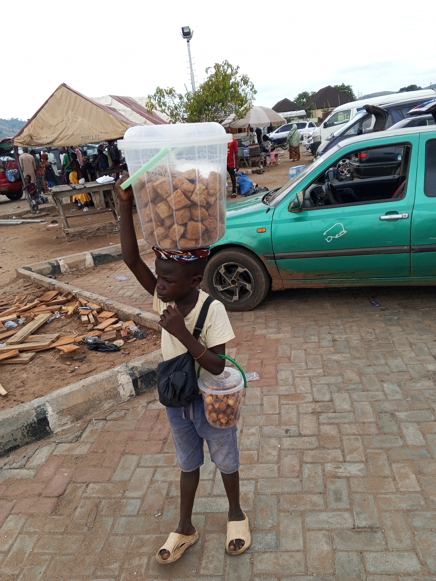 A young boy stands on a brick-paved area in Lokoja, Nigeria, with a large bucket of what appears to be fried dough on his head and a smaller bucket in his left hand. He wears a light-colored t-shirt, blue shorts, and sandals. A small black bag is slung across his chest. He appears to be eating something.
In the background, a green car is parked beside him. Further behind, there is a crowd of people gathered under a large tent, and several other cars are parked nearby. The environment is open and appears to be a market or gathering place. Pieces of wood and debris are scattered on the ground near a curb.