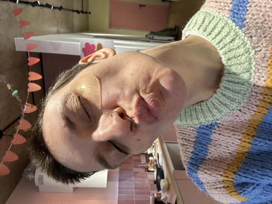 An individual is depicted in a medium close-up, indoors, presumed to be in a residence in Batumi, Georgia. The subject, with short dark hair, has their eyes closed and a gold-colored hydrogel under-eye patch applied beneath each eye. A silver septum ring and a silver nostril ring are visible on the nose. The lips are pursed in a "kissing" expression. The individual wears a thick-knit sweater featuring horizontal stripes in light blue, pink, yellow, and light green, over a white crewneck undershirt.
The background on the left reveals a kitchen area with light pink rectangular wall tiles and matching light pink base cabinets with white countertops. Several items, including a dark pan and various containers, are scattered on the counter. The background on the right features a white doorframe with a pink door partially open beyond it, revealing a darker interior. A light-colored garment or bag hangs near the doorframe. A multi-colored flower decoration is affixed to the white doorframe. Overhead, the ceiling appears to be exposed concrete. Pink heart-shaped garland is strung across the top of the frame, and a dark geometric garland hangs on the right side.