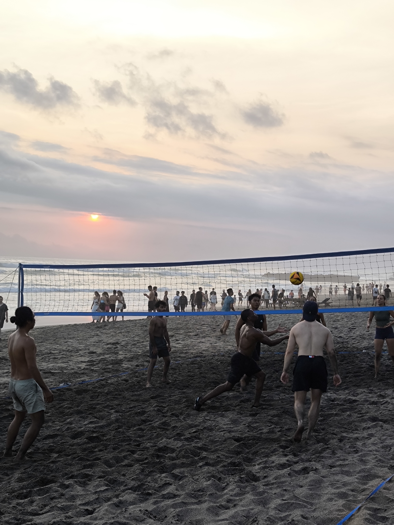 In North Kuta, Indonesia, on a beach at sunset, a group of people are playing volleyball. The sky is filled with soft, pastel clouds, and the sun is a bright orange disk near the horizon, casting a warm glow on the scene. The ocean stretches out behind the players, with gentle waves lapping the shore. The sand is dark and textured, with footprints and the lines of the volleyball court clearly visible. A net divides the players, who are in various stages of the game. Some are poised to hit the ball, others are running, and some are observing the play. The players are dressed in casual beach attire, mostly shorts and t-shirts. The atmosphere is lively and energetic, with the focus on the sport and the shared experience of a sunset game.