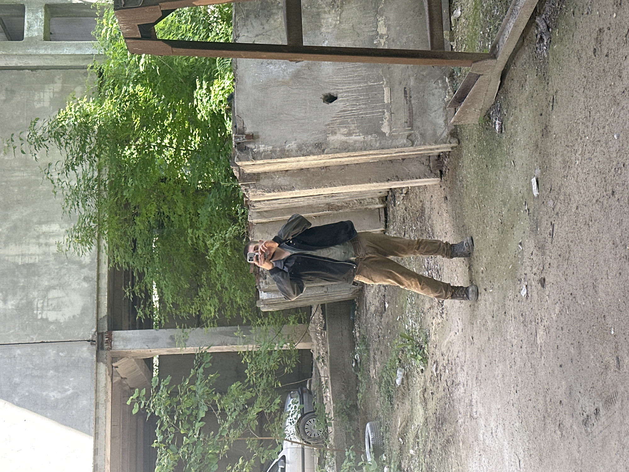 The image shows a man standing in an outdoor, industrial or construction site setting, holding a smartphone to take a photo of himself or his surroundings. The man is wearing a dark jacket, tan pants, and dark boots. He is positioned in the foreground, with a stack of large, concrete slabs to his right and a car visible in the background to his left. Lush green trees and bushes are present throughout the scene, suggesting an urban or semi-urban environment. The ground is covered in dirt and debris, and there are remnants of concrete structures, indicating a likely abandoned or disused area. The overall impression is one of decay and overgrowth, common in industrial zones or construction sites that have been left unfinished or have fallen into disrepair.
The location is identified as the former "Textil" factory in the city of Chișinău, Moldova. This site has a history of industrial activity and is now largely derelict, with areas being repurposed for commercial or residential use, while others remain abandoned. The presence of overgrown vegetation and discarded building materials is characteristic of such locations in Chișinău.