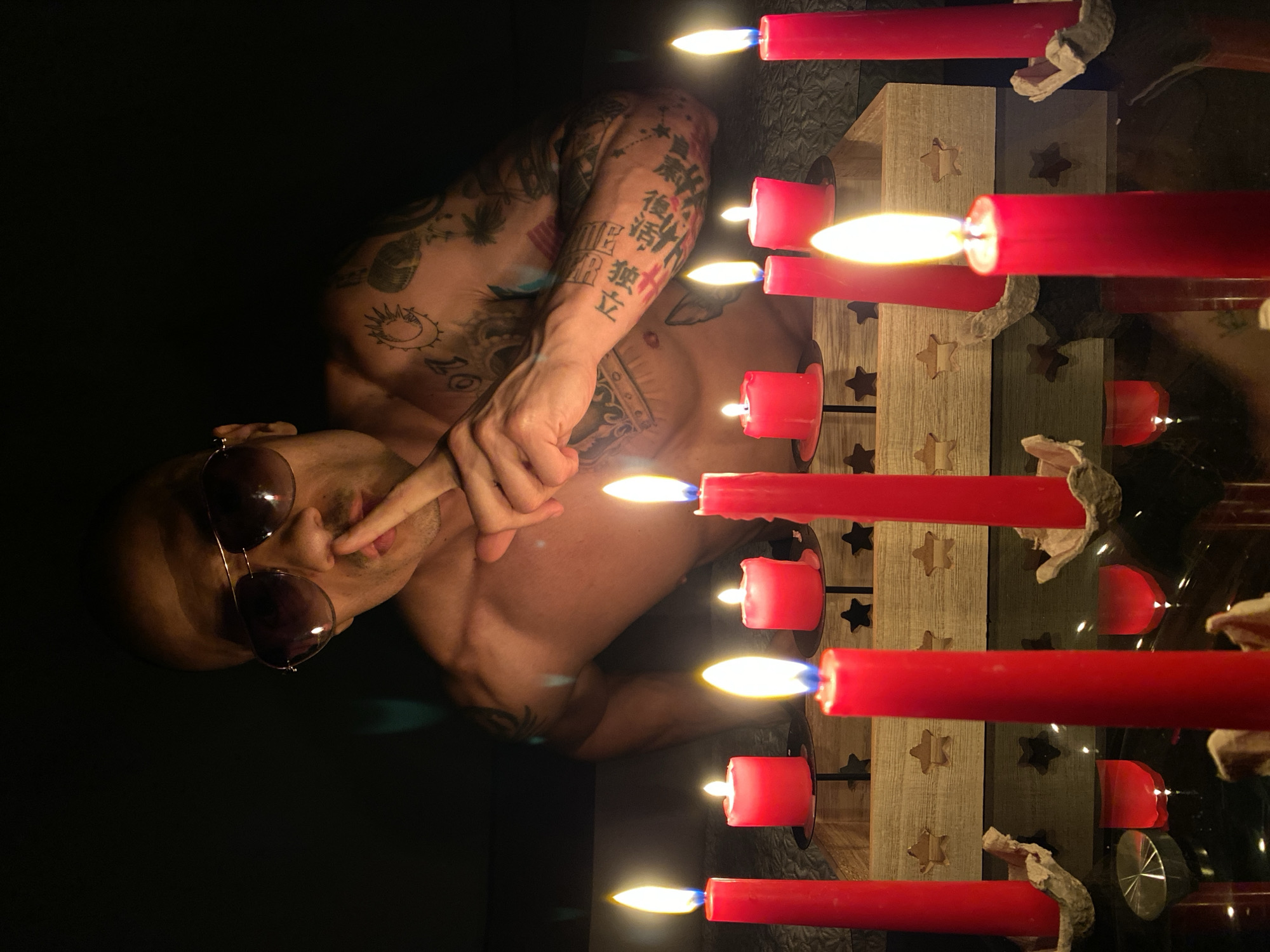 The image features a person, likely male, lying down with their torso visible, illuminated by the warm light of multiple red candles. The person is shirtless and heavily tattooed, with various designs covering their arms and chest, including a prominent sun and crescent moon motif. They wear dark, round sunglasses and have one hand raised, with a finger gently touching their lips, creating a contemplative or posed expression.
Several red pillar candles are prominently displayed, burning brightly and providing the main light source. These candles are set within a wooden holder featuring carved star cutouts, and the entire arrangement rests on a reflective surface, possibly glass, which shows reflections of the flames and candles. Some of the candles exhibit dripped wax, indicating they have been burning for a while.
The scene is set in a very dark indoor environment, suggesting it is night or a dimly lit room, emphasizing the glow of the candlelight. The overall atmosphere is intimate and somewhat mysterious.
Visible text on the person's right arm tattoos includes Japanese characters, such as "独立" (dokuritsu, meaning independence) and "後悔" (koukai, meaning regret).
This image was captured in Pleven, Bulgaria.