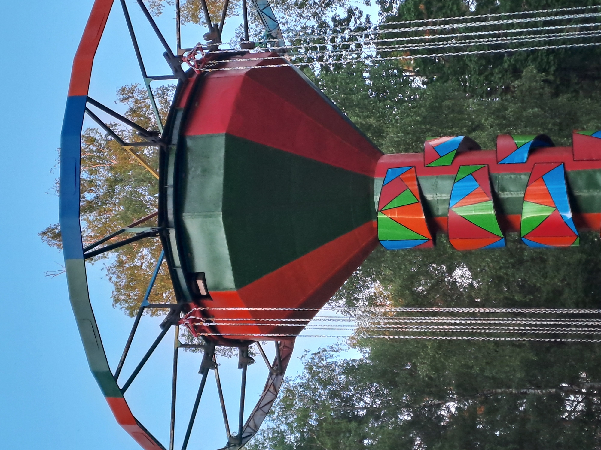 The image shows a fragment of a swing carousel against the backdrop of trees and the sky. The carousel has a large central cone-shaped structure painted in red and green. The individual swings hang from the central structure via chains. The seats of the swings are decorated with colorful geometric patterns. Metal support structures extend from the central part of the carousel, painted in blue, green, and red. The background features trees with green foliage and a blue sky. There is no evidence to suggest that the image was taken in Noginsk, Russia.