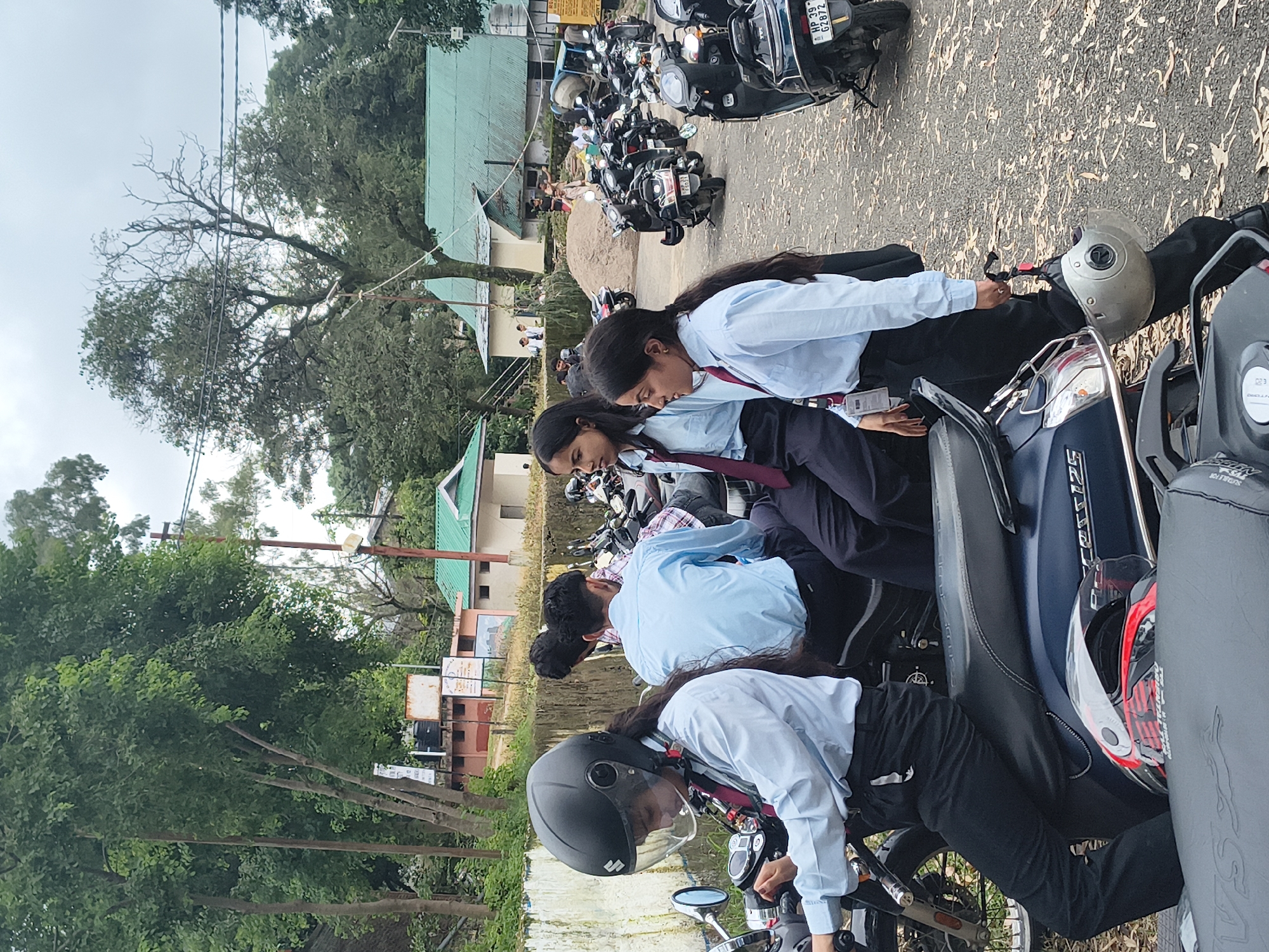 The image depicts a group of young individuals, likely students, standing and sitting on scooters in an outdoor setting. The students are dressed in light blue shirts and dark trousers, with some wearing ties. They appear to be in a parking lot or a similar area, possibly on a campus or public space. Multiple scooters are parked around them. In the background, there are buildings with green roofs and trees, suggesting a semi-urban or rural environment. The presence of a license plate visible on one of the scooters, "HP 39 G2872," indicates the location is Himachal Pradesh, India. Given the architectural style of the buildings and the lush greenery, this scene is consistent with the city of Dharamshala, India, known for its hilly terrain and institutional buildings.