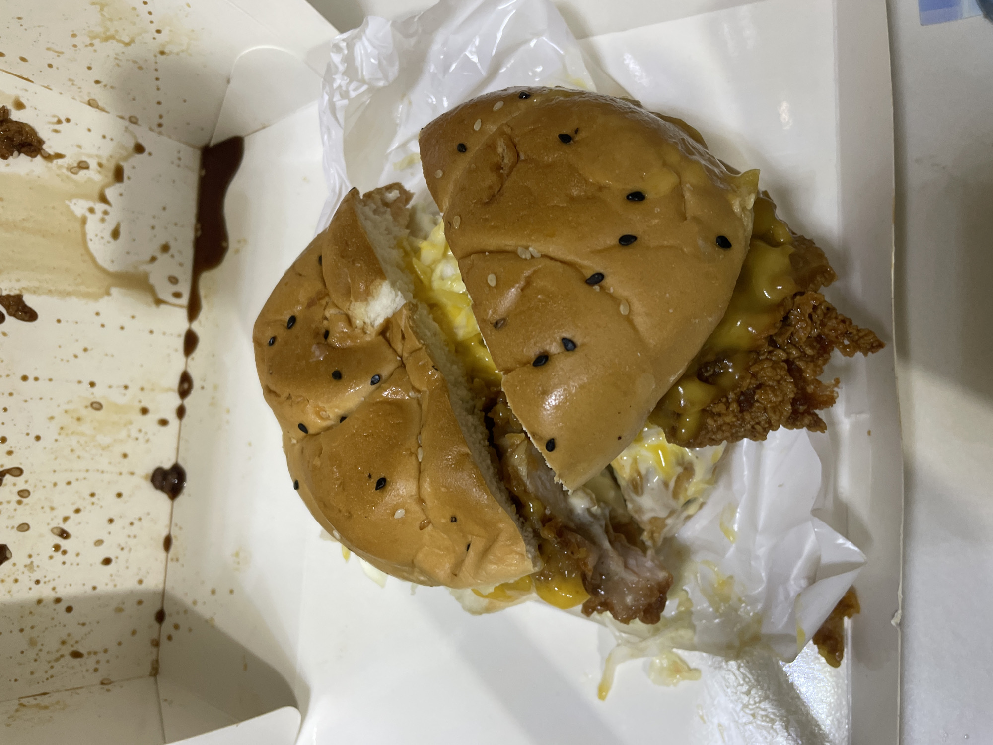 The image shows a partially eaten burger inside a white takeaway box. The burger is cut in half, with the two halves slightly separated. The bun is topped with black sesame seeds and appears slightly greasy. The filling includes fried chicken, cheese, and a white sauce. The inside of the box is stained with brown sauce splatters. The burger rests on a crumpled piece of white parchment paper. The location is Davao City, Philippines.