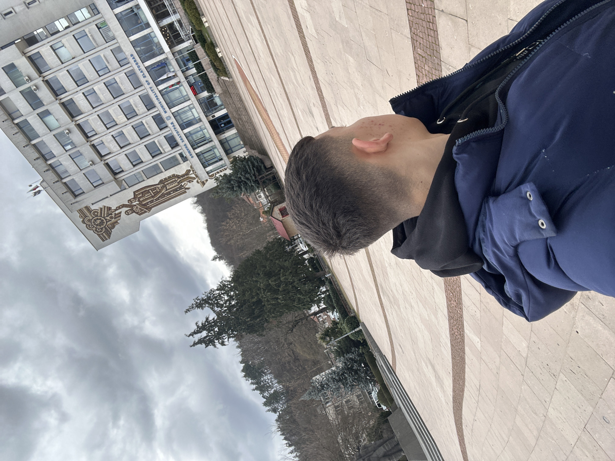 A medium, close-up shot taken from a high angle depicts the back of a person's head and the upper part of their body, clad in a dark hoodie and a blue puffer jacket. The person appears to be standing on a tiled pavement with alternating wide and narrow beige tiles that converge towards the top of the frame, creating a sense of perspective.
In the background, slightly off to the left, stands a multi-story building with a repetitive pattern of windows. The building displays the inscription "Община Златоград / Municipality of Zlatograd" in Bulgarian, indicating its location in Zlatograd, Bulgaria. A decorative mosaic is visible on the lower part of the building's façade.
Beyond the building, the landscape reveals a scene of trees and a hillside shrouded in cloudy, overcast weather. The general environment suggests an urban or semi-urban setting with a mix of architectural and natural elements under dull lighting conditions. The scene is still.