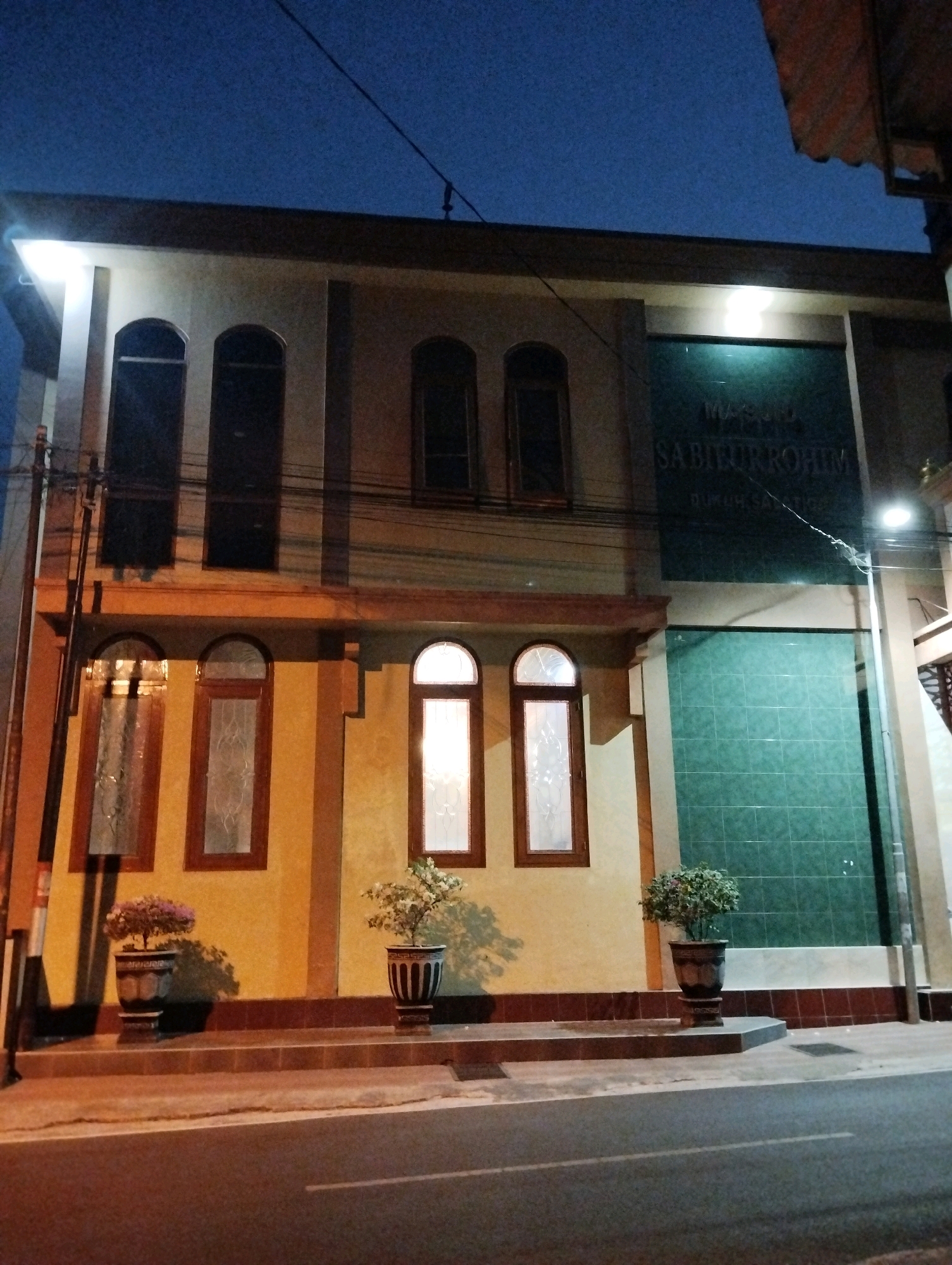 The image depicts a building at dusk or night in the city of Salatiga, Indonesia. The building appears to be a mosque, as indicated by the partially visible text on its facade: "MASJID SABILURROHIM." The building has a light-colored facade with arched windows on the upper level and rectangular windows with ornate glass on the lower level. Two decorative potted plants are placed on steps leading to the entrance of the building. A dark green tiled section is visible on the upper right side of the building, possibly part of its facade or an adjoining wall. The scene is lit by artificial lights, creating shadows and highlighting the building's architecture. A street is in the foreground, indicating an urban setting. The sky is dark blue, typical of twilight or nighttime. No people are visible in the image.