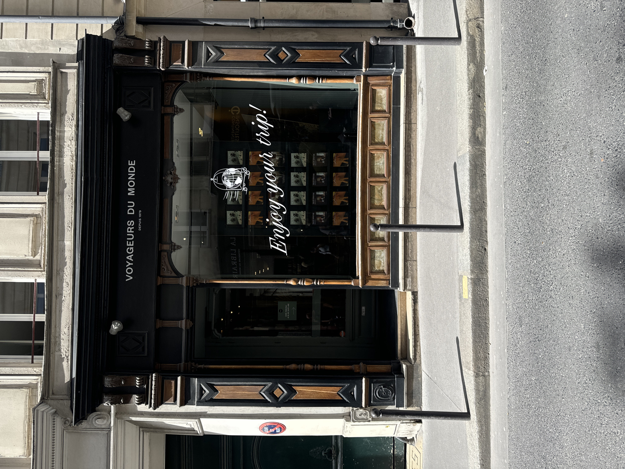 This media file captures the detailed facade of a shopfront in Paris, France, during daytime under clear, sunny weather. The building displays classic Parisian architecture with light-colored stone or stucco walls, featuring ornate moldings and window frames.
The shopfront itself is elaborately designed with dark wood and decorative elements. A prominent black signboard above the large glass display window reads "VOYAGEURS DU MONDE" (Travelers of the World) in white capital letters, with "DEPUIS 1979" (Since 1979) beneath it. A white security camera is mounted just below this sign on the left.
The shop window is the focal point, featuring a large white logo depicting a globe within a birdcage-like structure on the left side. To its right, in elegant white cursive, is the phrase "Enjoy your trip!". Below this, the window displays multiple square panels, likely showcasing images of various travel destinations or packages, some appearing to feature palm trees. Reflections in the window also show parts of the interior and the reversed text "LA LIBRAIRIE" (The Bookstore/Library).
The lower section of the storefront incorporates intricate dark wooden panels with diamond patterns and additional carved details. To the far left, a red circular "NO PARKING" sign is partially visible on the building's base.
The scene includes the sidewalk in front of the shop, lined with dark metal bollards, suggesting a pedestrian area or restricted parking zone. Beyond the sidewalk, a paved street or road is visible. No people are seen in the image.