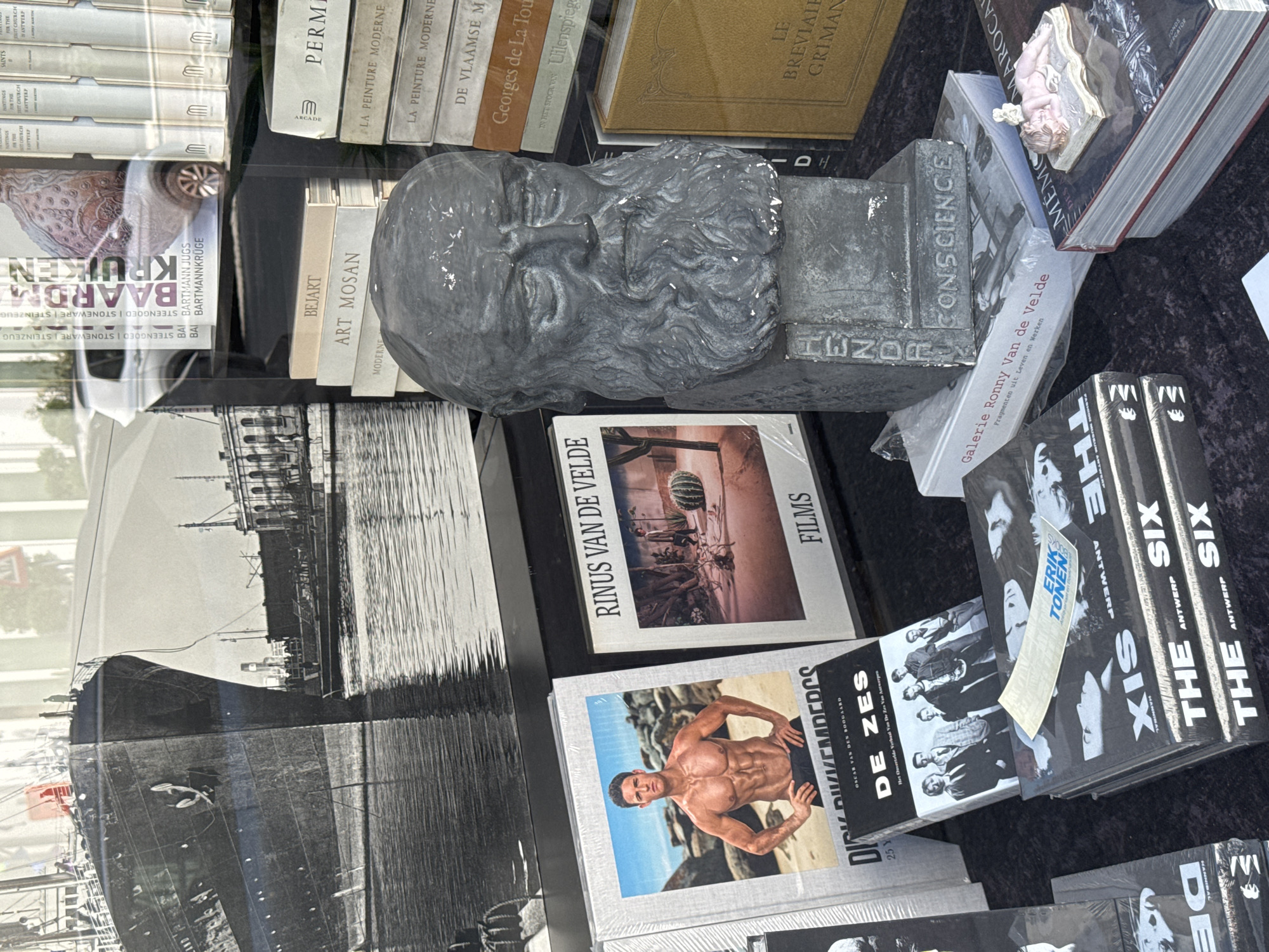 The image depicts a display of books and a bust in what appears to be a shop window or outdoor market stall. The overall scene is a close-up shot focusing on the items for sale.
Dominating the center of the image is a grey stone bust of a man with a long beard and intense eyes. The bust is placed on top of several books. The base of the bust has text carved into it, reading "HENDRICKX" on one side and "CONSCIENCE" on another.
In front of the bust, on the left side of the frame, is a large, open book with a photograph of a muscular man on the cover. The text on this book is partially obscured but includes "DIRK DIJKKEMPERDS". Next to it, to the right, is another book titled "RINUS VAN DE VELDE FILMS" featuring a photograph of a desert landscape with a cactus and two figures.
To the right of the bust, a stack of books is visible. One prominently displays the title "LE BREVIAIRE GRIMANI" in gold lettering on a yellow cover. Above this, other books with titles like "LA PEINTURE MODERNE" and "DE VLAAMSE MEESTERS" can be discerned.
At the bottom of the frame, a stack of books with black covers is arranged. The titles "THE SIX" and "ANTWERP" are repeatedly visible on these books. A sticker with "ERIK TONEN" is also present on one of these books.
In the background, through a reflective surface that acts as a window, a black and white photograph of a large ship docked at a harbor is visible. Beyond the ship, some buildings and a silver car are also faintly discernible, suggesting an urban setting. The reflection indicates the display is likely inside a shop with a view of the street or an adjacent area.
The context of Antwerp, Belgium is suggested by the presence of the word "ANTWERP" on the books, and the general aesthetic of a European city displaying cultural items. Without specific landmarks or signage directly linking to a known location within Antwerp, the precise spot cannot be identified beyond being a commercial display in the city.