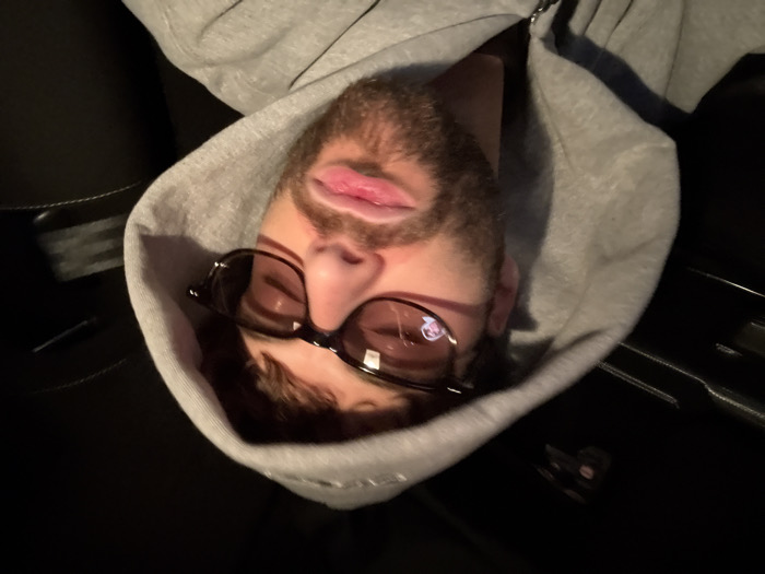 The image displays a close-up portrait of an adult male situated inside a vehicle cabin, in Givors, France. The subject, positioned centrally, is wearing a grey hooded sweatshirt with the hood pulled over his head. He has short, dark brown hair and a beard. He is wearing dark-framed eyeglasses with amber-tinted lenses. A distinct, miniature reflection of a human face is visible within the upper right lens of his eyeglasses. His lips are slightly parted. The background is uniformly dark and indistinct, indicating the interior of a vehicle with dark upholstery and headliner elements. The lighting is dim, illuminating the subject's face from the front.