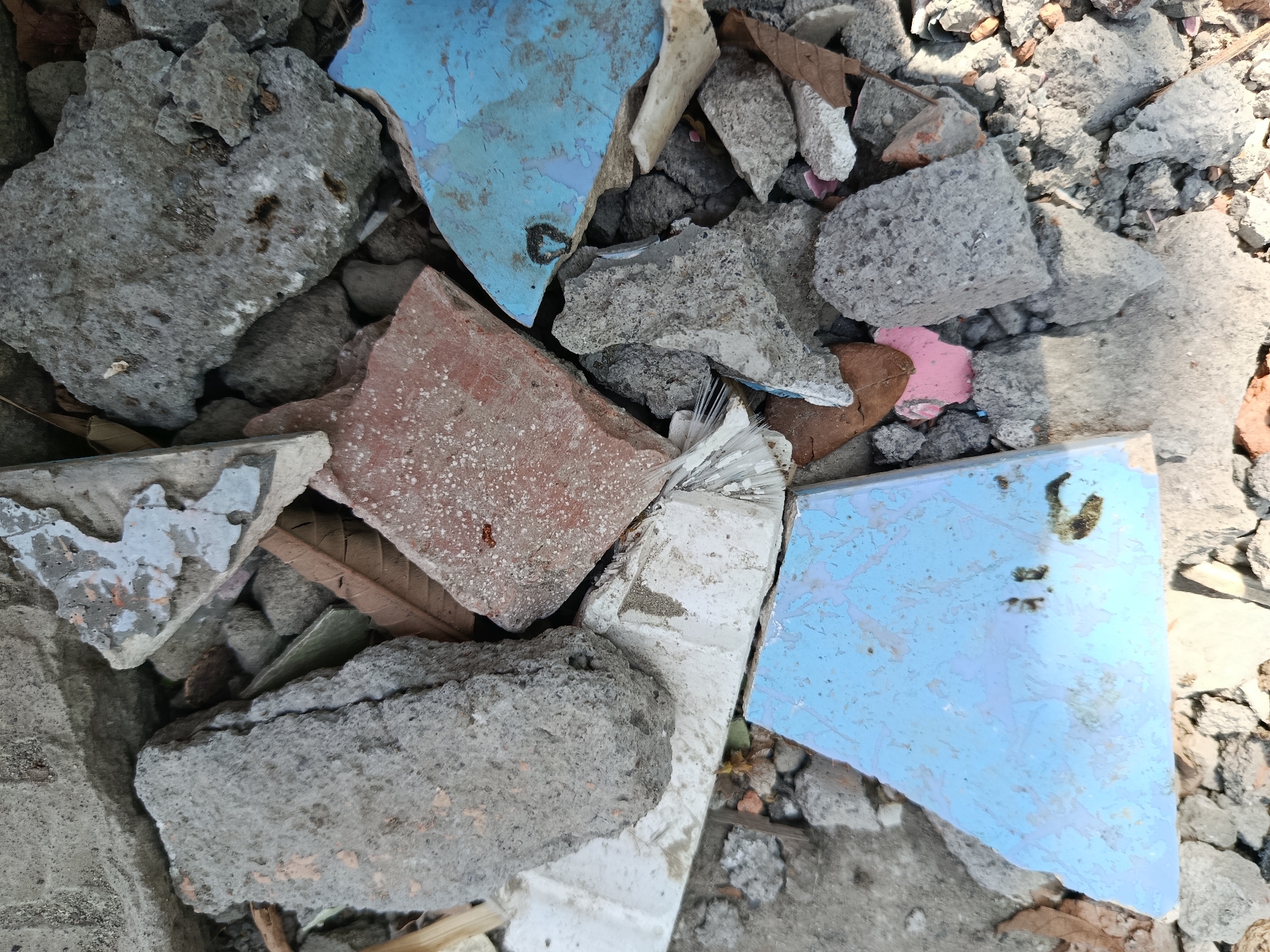 The image shows a pile of rubble, likely from a demolition or construction site, in Wanasari, Indonesia. The debris consists of broken concrete, bricks, and shattered tiles in various colors including blue, pink, and red. Some pieces of tile show signs of wear and tear, with peeling paint or faded patterns. Dried leaves are scattered amongst the rubble, suggesting an outdoor setting. The lighting indicates it is daytime, with shadows cast by the debris. There are no people or animals visible in the image. The scene appears to be a common sight in areas undergoing development or renovation.