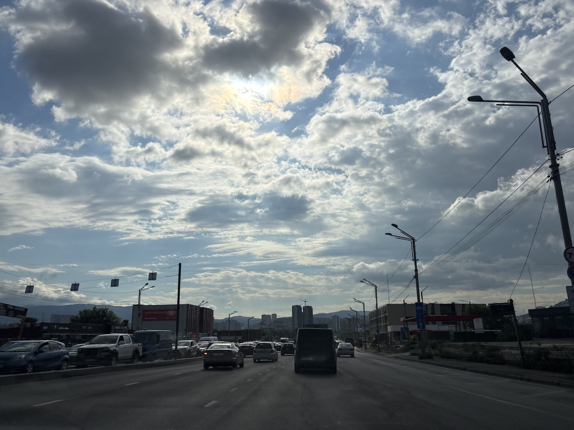 The image depicts a street scene in Tbilisi, Georgia, likely on a cloudy day. The perspective is from inside a vehicle, looking forward along a multi-lane road congested with traffic. Cars, vans, and trucks are visible in various lanes. On the right side of the road, there are streetlights and utility poles with wires crisscrossing overhead. Buildings line the right side of the road, some appearing to be under construction or recently built, with one featuring red and white signage. In the background, a cityscape with high-rise buildings and cranes is visible, with mountains forming the distant horizon. The sky is filled with a mix of dark and light clouds, with the sun partially obscured, creating a diffused light. Traffic lights and road signs are also present, though not clearly legible.