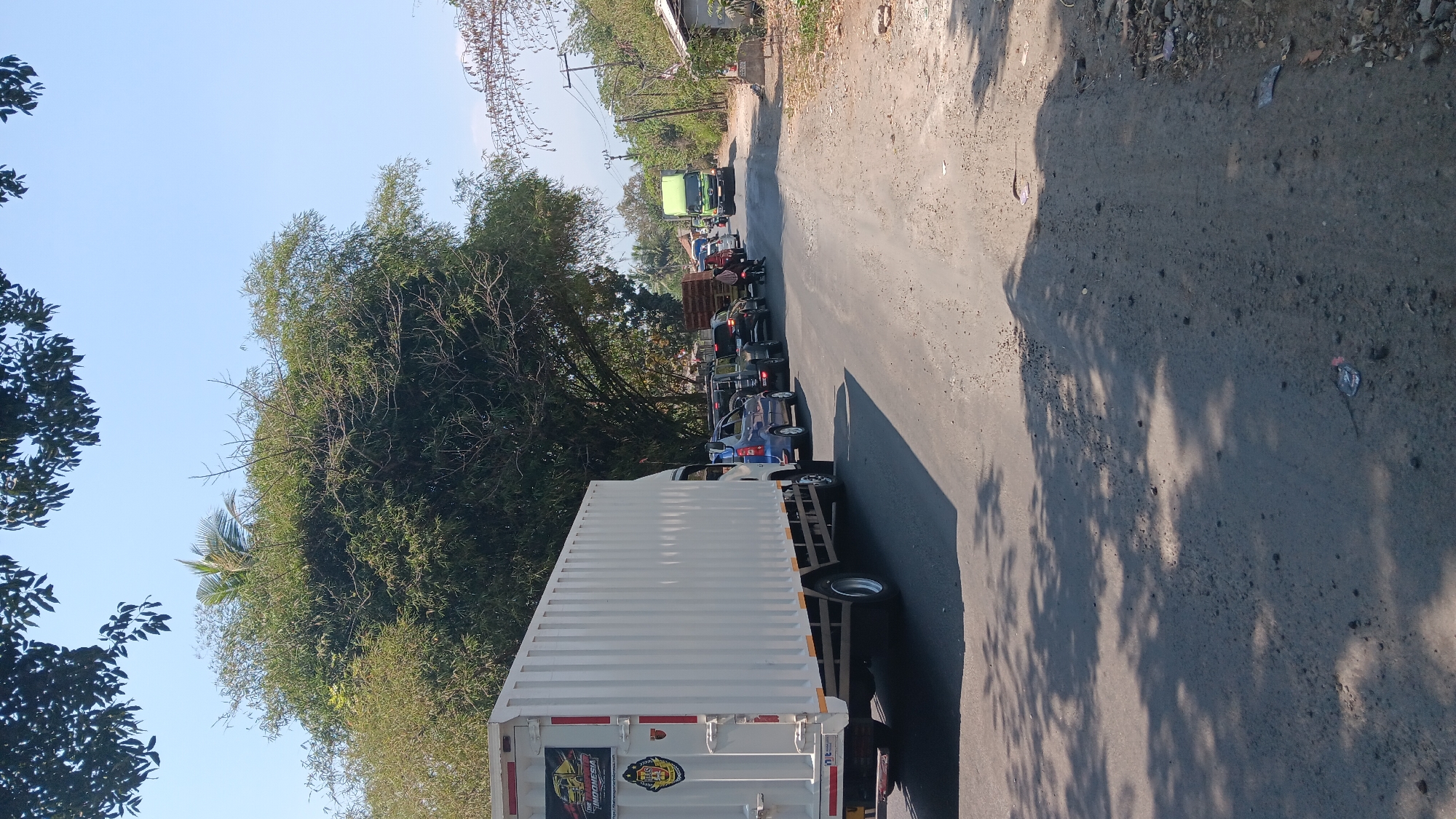 The image depicts a traffic jam on a rural road in Jonggol, Indonesia. In the foreground, a large white truck is parked on the left side of the asphalt road. Several vehicles, including a blue sedan, SUVs, and motorcycles, are stopped behind it, forming a queue. The road is lined with trees on both sides, with foliage creating dappled shadows on the pavement. In the background, a green truck is visible further down the road, also part of the traffic. The sky is clear and blue, indicating a sunny day. The surrounding environment appears to be a semi-rural area, with some houses and vegetation visible on the right side of the road. The overall scene suggests a delay or congestion on a journey.