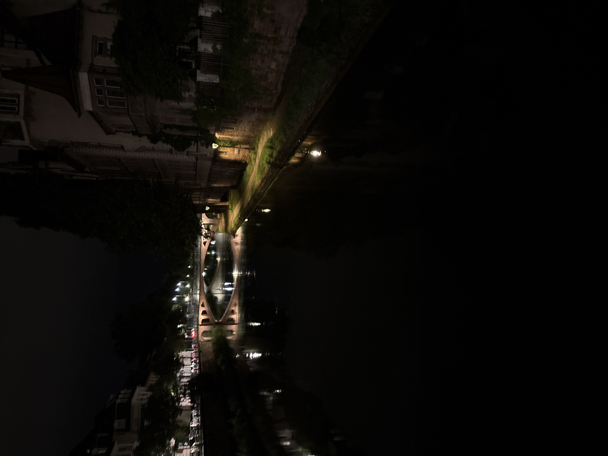 The image shows a night scene along the canals of Strasbourg, France. The view is of a canal with buildings lining the waterway. The structures consist of several floors, the architectural style is traditional. Light sources are primarily from streetlights along the bank and reflections in the water. The area around the canal is vegetated. A bridge spans the canal further down.