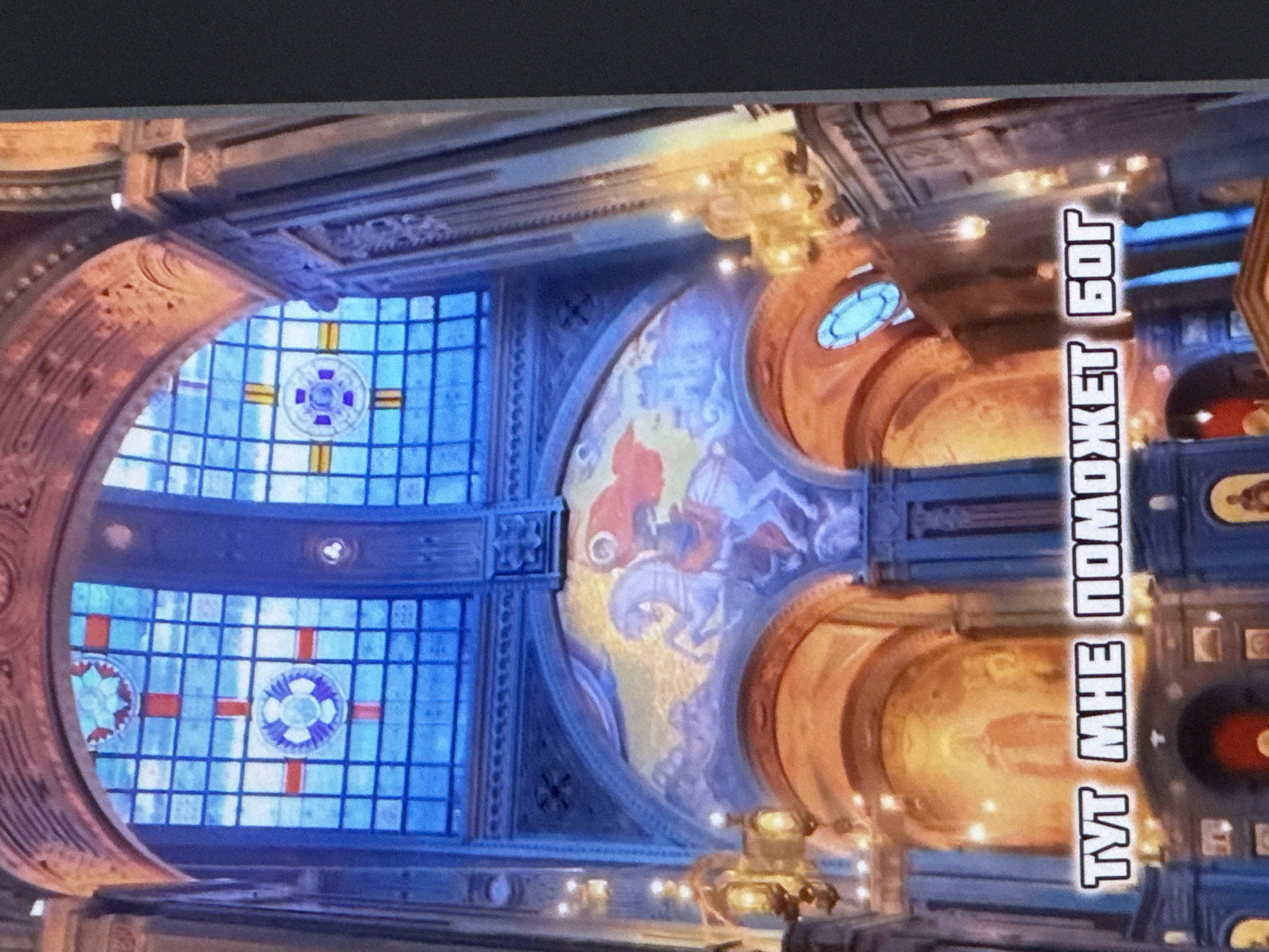 This media file captures the interior of a grand, ornate building, likely a church or cathedral, given the architectural style and the presence of stained glass windows and religious artwork. The scene is illuminated by artificial lights, suggesting it is either nighttime or the interior is dimly lit.
The prominent subjects are the architectural elements of the building. We see large, arched stained glass windows with intricate geometric patterns and circular motifs featuring crosses. Above the windows, ornate molding and decorative carvings adorn the walls and ceiling. A large mural depicting figures, possibly religious in nature, is visible on an arched section of the wall. The ceiling features chandeliers, adding to the grandeur of the space.
A striking detail is the vertical text written in a bold, white font with a black outline. The text reads "ТУТ МНЕ ПОМОЖЕТ БОГ" which translates from Russian to "GOD WILL HELP ME HERE". This text is positioned on the right side of the image, oriented vertically.
The overall impression is one of solemnity and grandeur. The lighting creates a dramatic effect, highlighting the textures and colors of the architecture and artwork. The location cues, such as the religious iconography and the architectural style, strongly suggest a religious setting. While no specific time of day or weather is discernible, the interior lighting implies the activity is taking place within the building, regardless of external conditions. The presence of the text "GOD WILL HELP ME HERE" conveys a sense of faith and hope associated with the location.