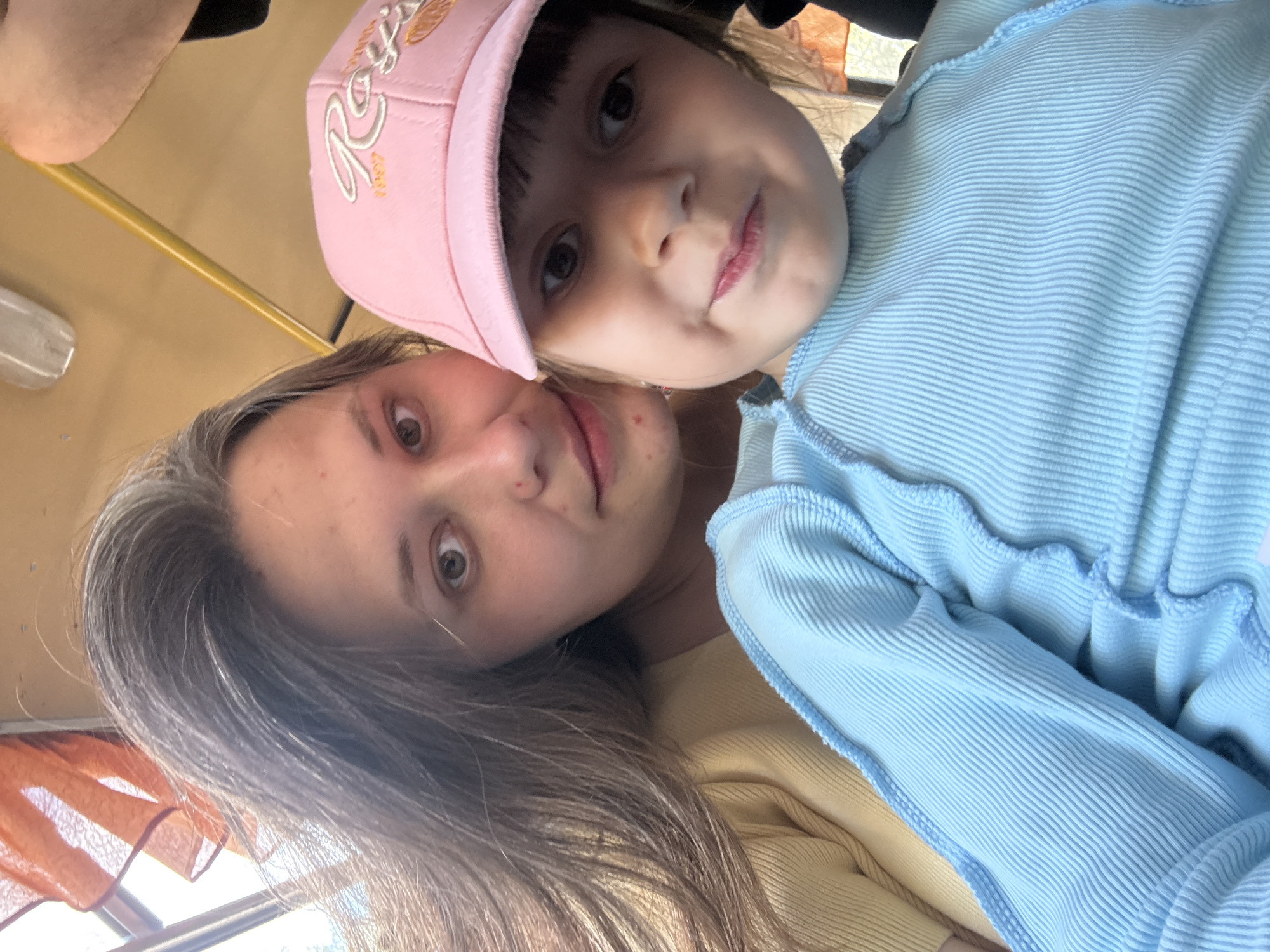 Two individuals, a young girl and a young woman, are captured in a close-up photograph. The girl, wearing a pink baseball cap with an embroidered design, is positioned closer to the camera. The young woman is slightly behind her. Both appear to be indoors. The girl is wearing a light blue ribbed top. The young woman is wearing a yellow top. The background is obscured but features an orange-toned curtain and a window. The image was captured in the city of Chaikovsky, Russia.
