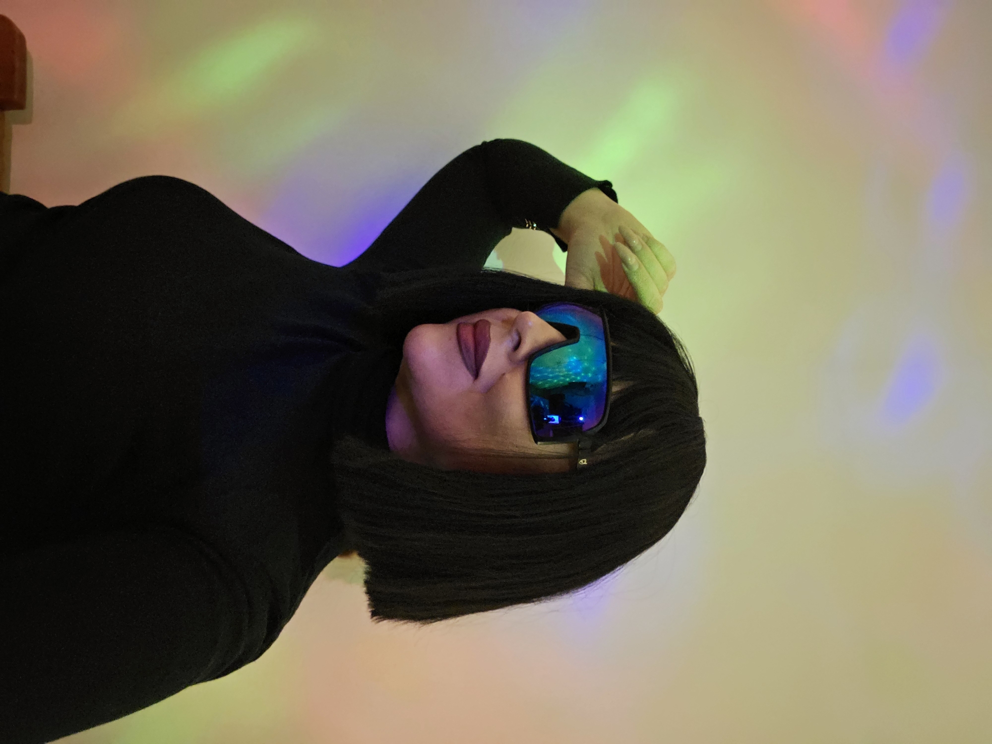 The image features a close-up portrait of a woman posing indoors. She is seen from the chest up, with her head slightly tilted upwards and to the side, and her right arm bent with her hand resting on her head. She has short, dark, straight hair, likely a bob cut. Her face is partly obscured by large, futuristic mirrored sunglasses that reflect vibrant blue, green, and purple hues, indicating the presence of colorful lights in the environment. Dark lipstick is visible on her lips beneath the sunglasses. She is wearing a black turtleneck shirt. Her extended fingernails, which are long and light-colored, are visible on her raised hand.
The background consists of a plain, light-colored wall, likely white or off-white, illuminated by various colored light projections. These lights cast distinct streaks and patches of red, green, and purple/blue across the wall, creating a dynamic and artistic backdrop. A small, indistinct reddish-brown object or wooden structure is partially visible in the upper left corner of the background. The overall scene suggests a stylized photoshoot or a fashion-oriented portrait, with the colorful lighting contributing to a modern and slightly mysterious aesthetic. No visible text is present in the image.