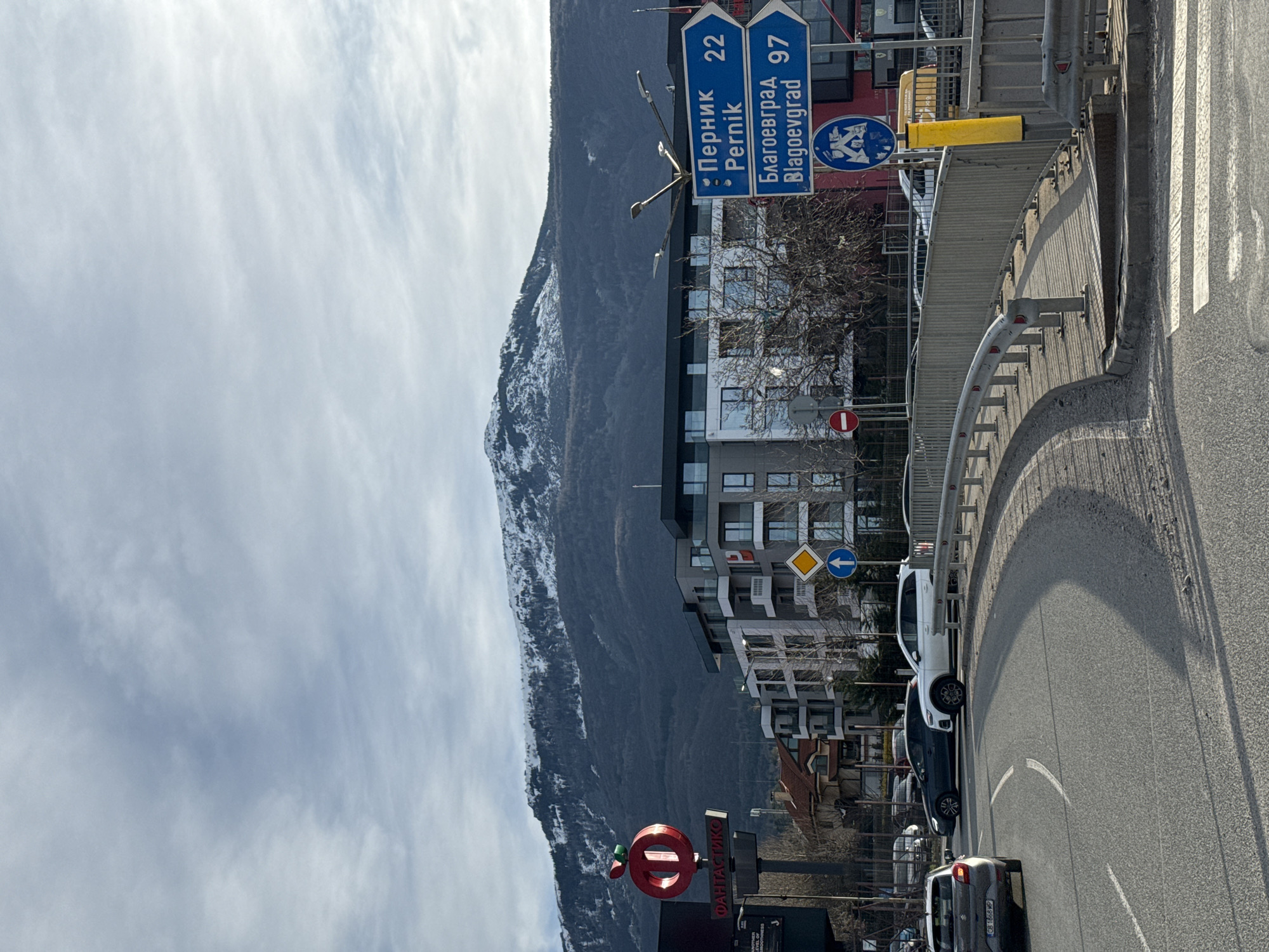 The image shows a street scene in Sofia, Bulgaria. A mountain covered in snow rises in the background, partially obscured by clouds. A road curves to the right, lined with a metal guardrail. Cars are driving along the road. A sign indicates that Pernik is 22 kilometers away and Blagoevgrad is 97 kilometers away. There is a large red sign with the word "ФАНТАСТИКО" (Fantastico) below it, indicating a supermarket. Buildings are visible behind the road. A pedestrian crosswalk is on the right side of the frame. A street sign indicates a turning road to the left.