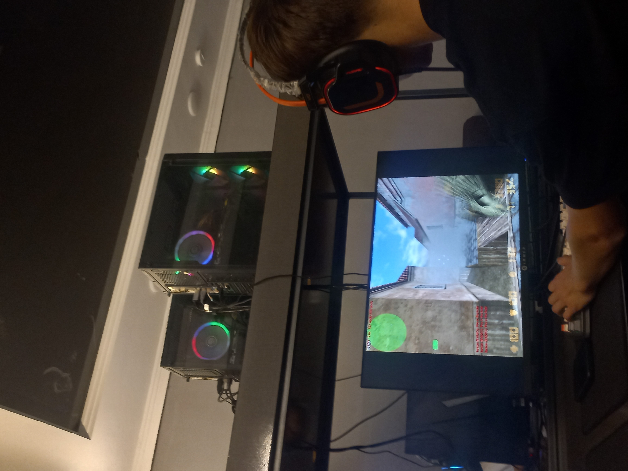 A person, seen from the side and slightly from behind, is wearing gaming headphones and playing a video game on a computer. The person has short brown hair and is wearing a dark t-shirt. They are looking at a computer monitor that displays a first-person shooter game with a cityscape background. The game appears to be Counter-Strike: Global Offensive, indicated by the in-game HUD which shows player information, a minimap, and text messages. The scene is set in what appears to be a home or gaming setup, with two desktop computer towers visible, each featuring RGB lighting on their fans. One computer is stacked above the other. The lighting in the room is dim, suggesting it might be evening or nighttime. The image is taken from a low angle, with the computer towers and monitor taking up a significant portion of the frame. The wall behind the setup is painted a light color and has moldings.