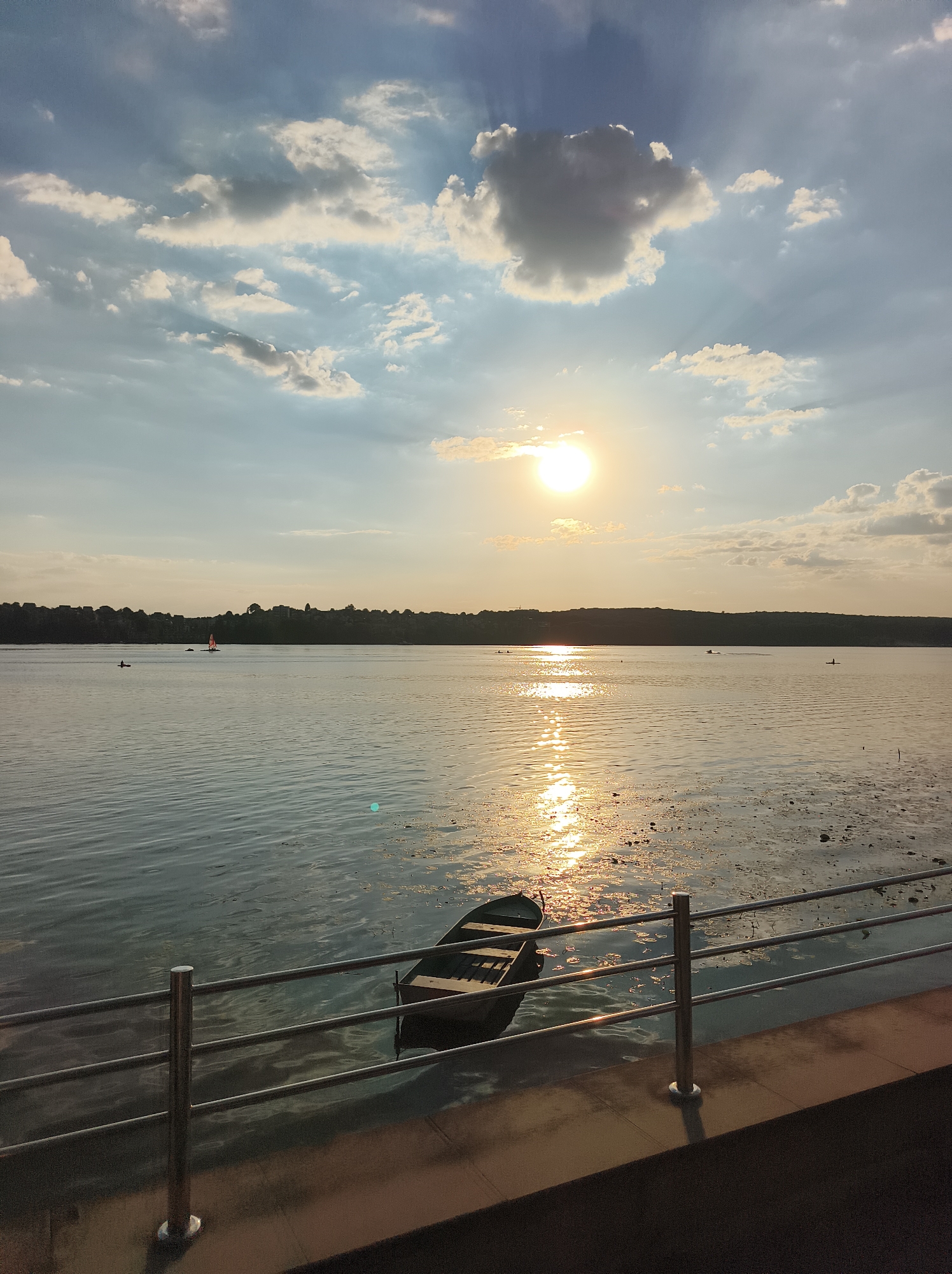 The image depicts a serene sunset over Ternopil Lake in Ternopil, Ukraine. The sun, a bright orb in the sky, casts a warm glow on the water, creating a shimmering reflection. Several clouds, some dark and others illuminated by the sun's rays, dot the sky.
In the foreground, a metal railing separates a paved walkway from the lake. Moored near the walkway is a small, dark-colored boat. The lake is calm, with gentle ripples on its surface. In the distance, a tree-lined shore forms the horizon. Several small figures can be seen on the water, including a sailboat and what appear to be individuals in kayaks or canoes. The overall atmosphere is peaceful and tranquil, capturing the beauty of a sunset by the water.